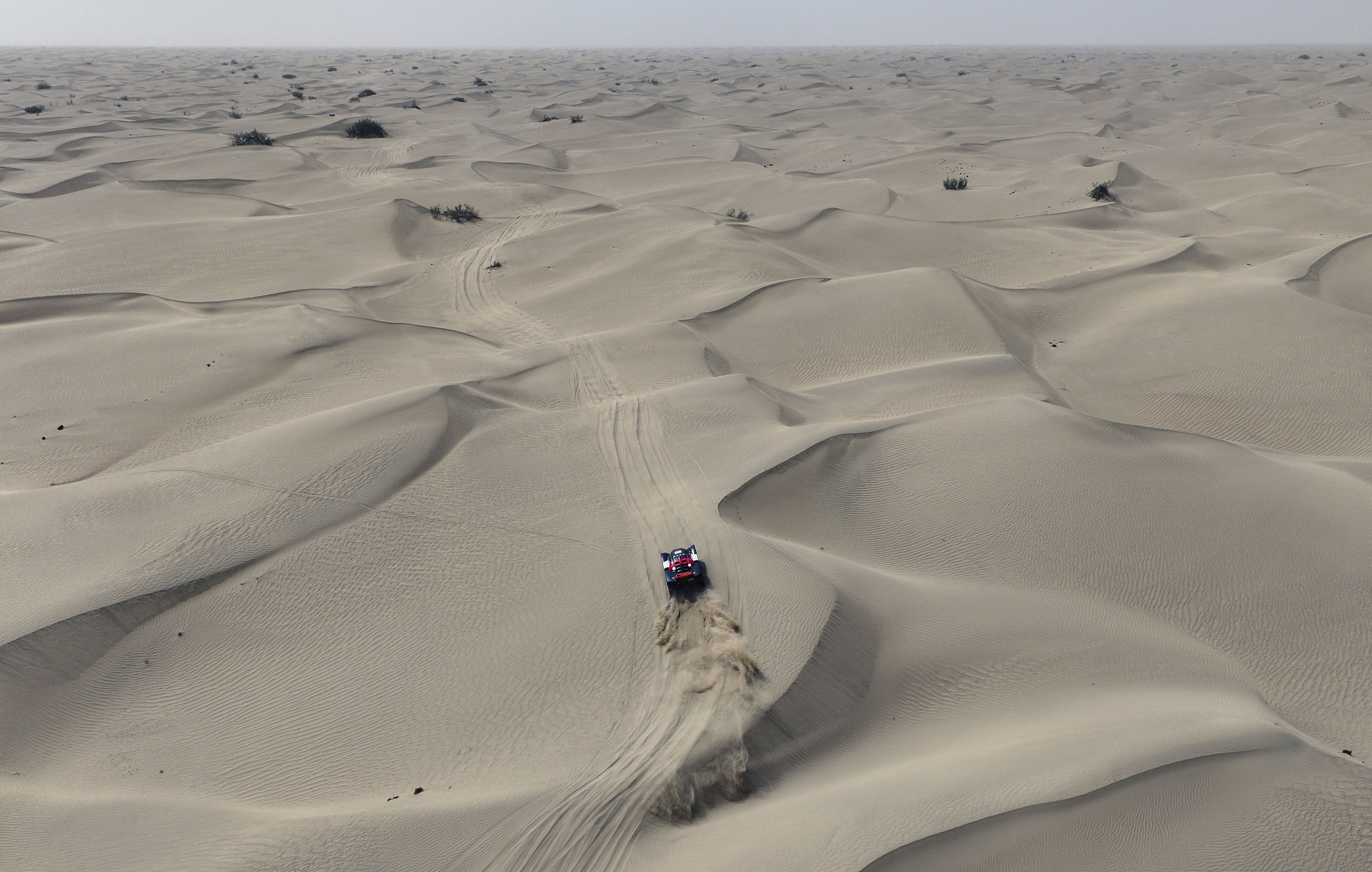 An aerial view of a rally race car driving across a broad field of sand dunes