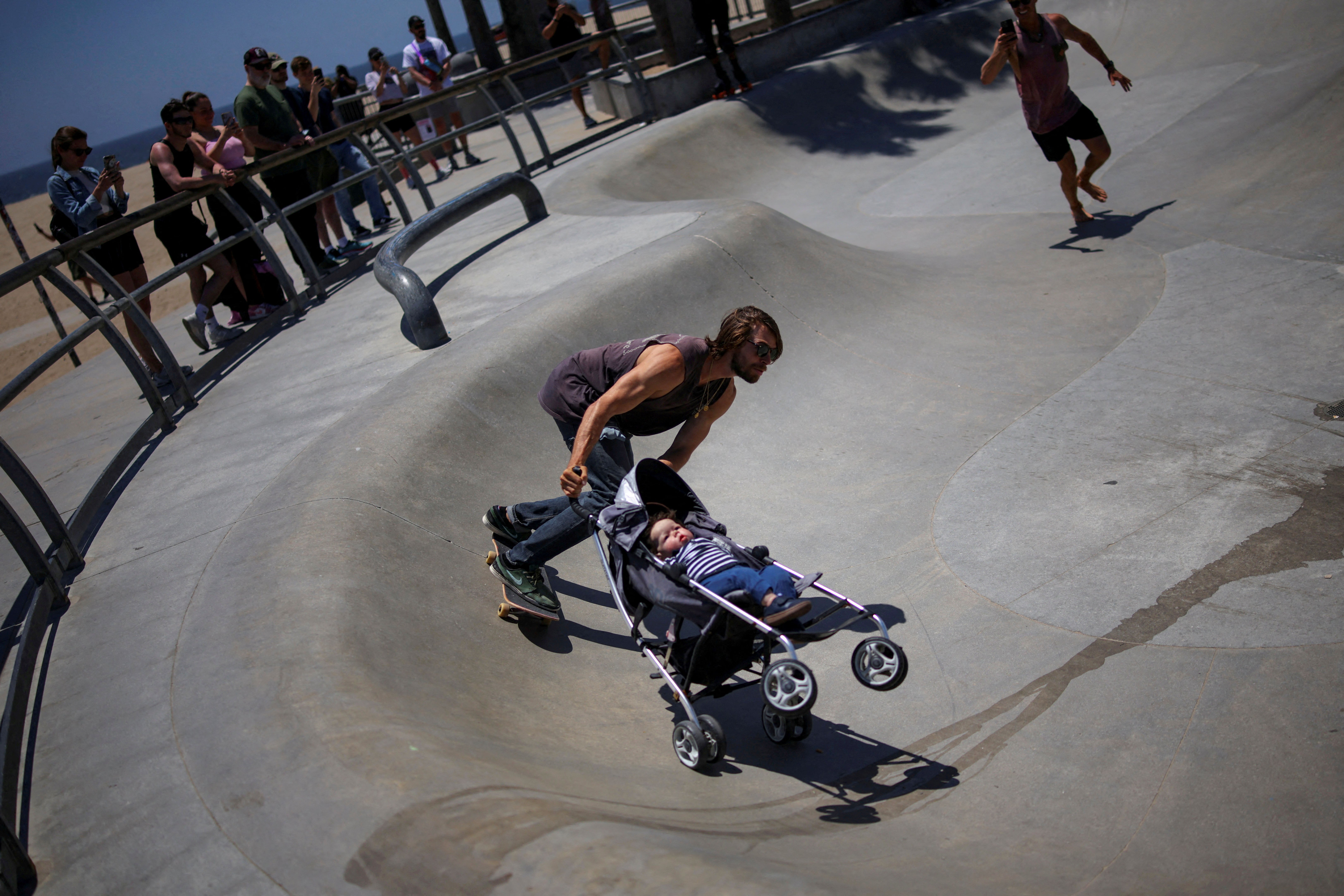 A skateboarder pushes a stroller with a doll as he rides through a skate park.