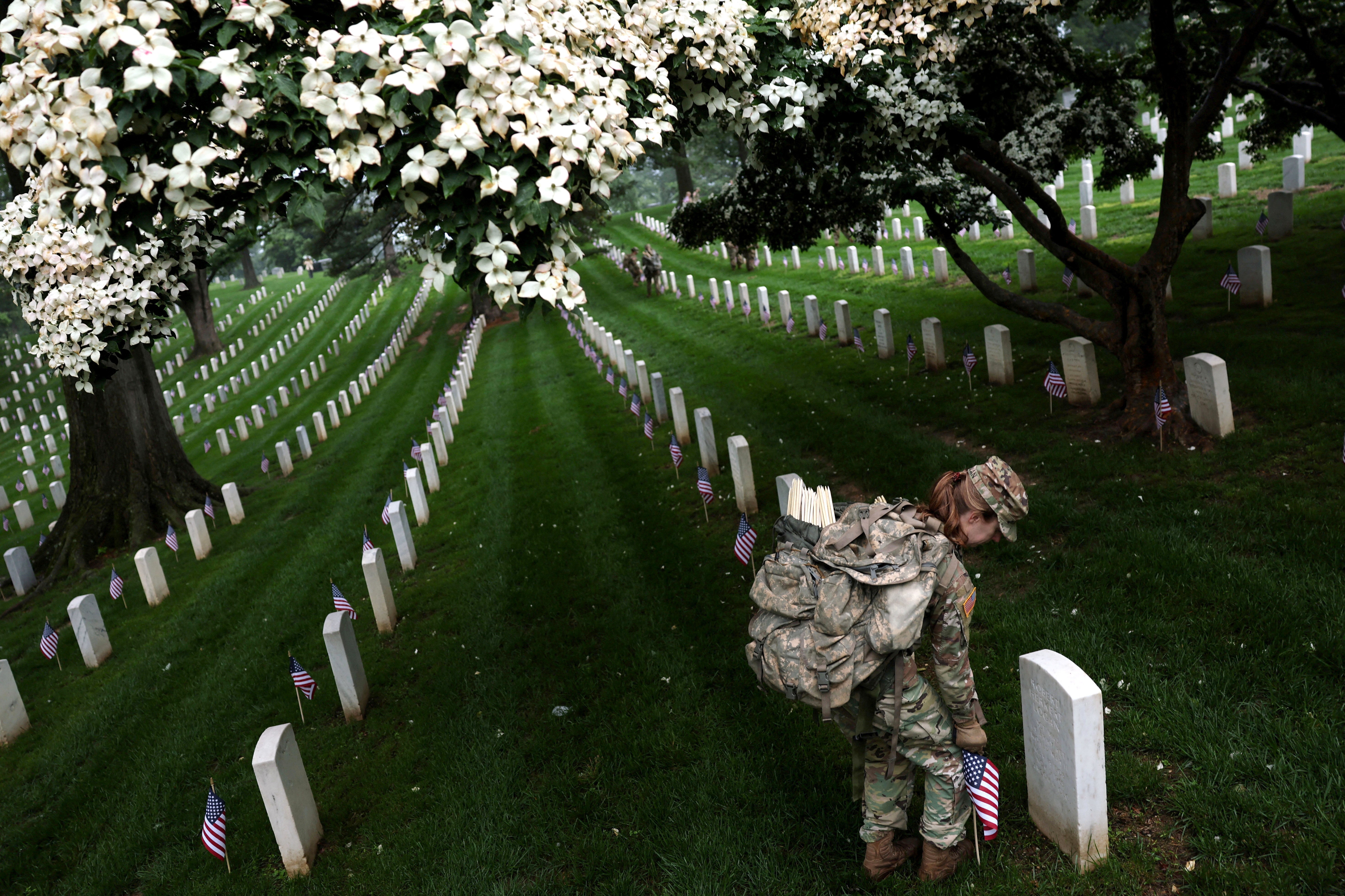 A soldier places a small American flag beside one of many gravestones standing in rows in a cemetery.