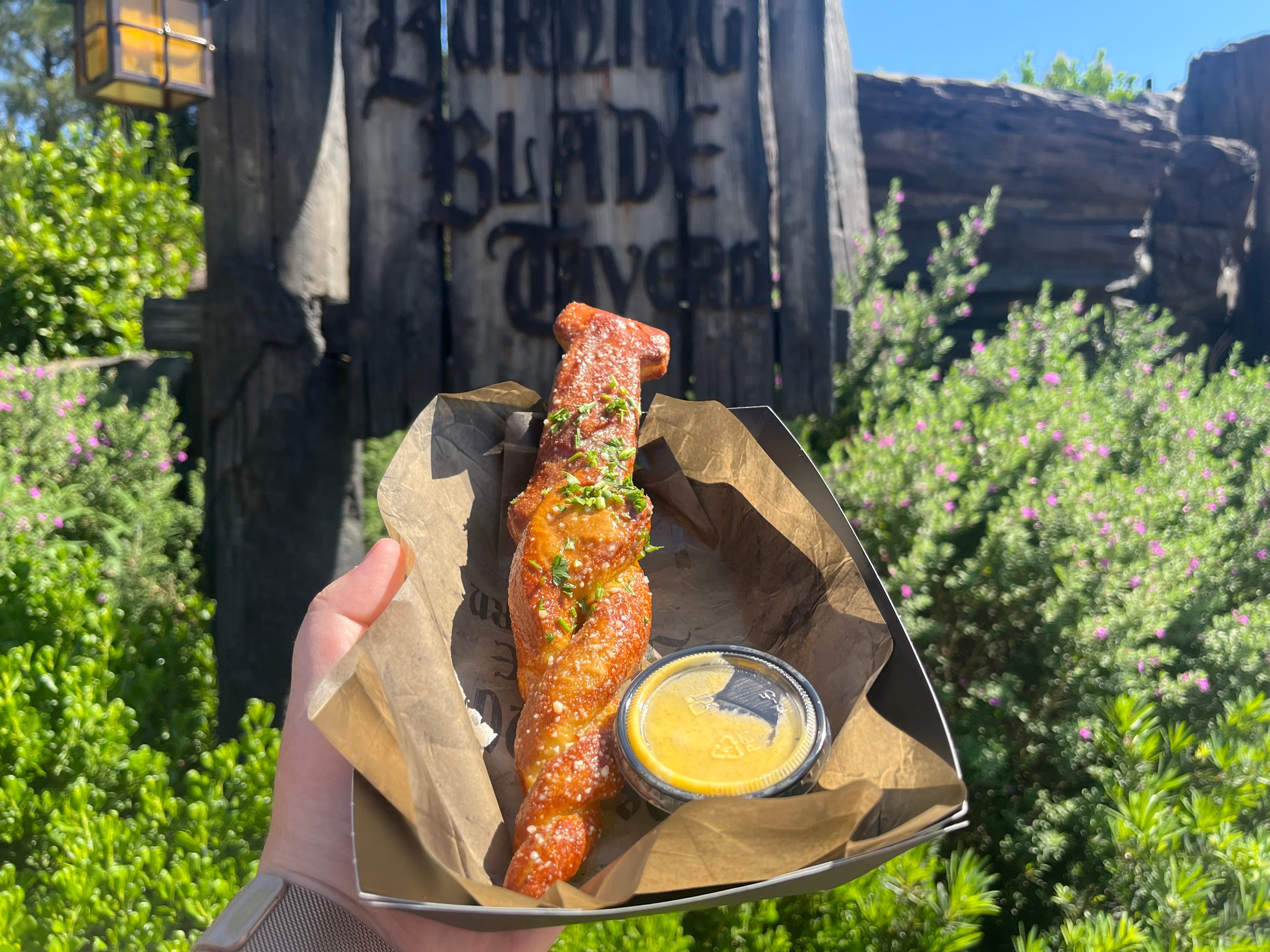 Pretzel shaped like a sword next to dipping cup of cheese on parchment paper being held in front of Burning Blade Tavern sign