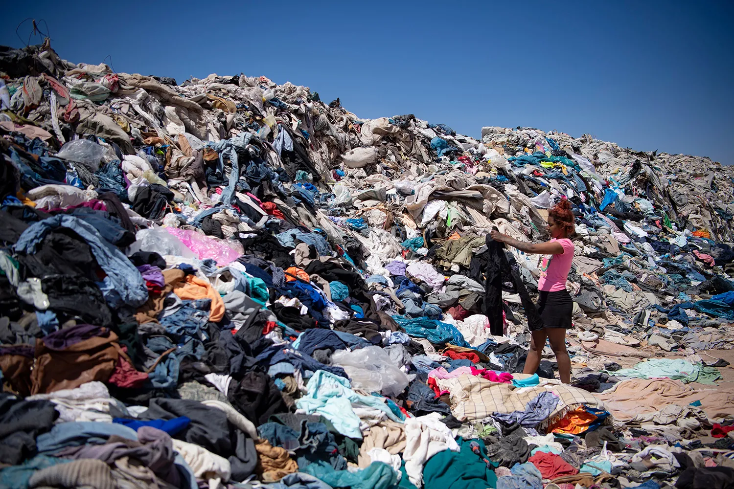 A woman in a pink shirt holds up an item of clothing, surround by a mountain of discarded clothes.