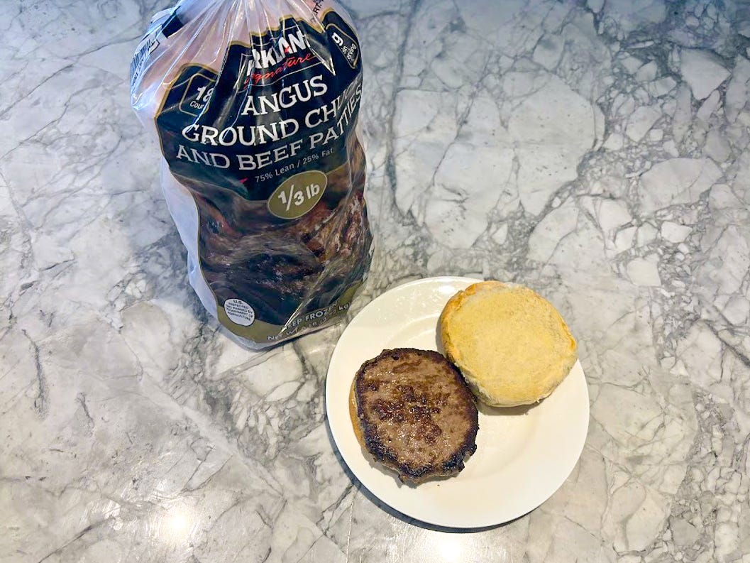 A package with a black label and an image of a grilled burger next to a white plate with a cooked burger on a bun with the top of the bun placed on the side
