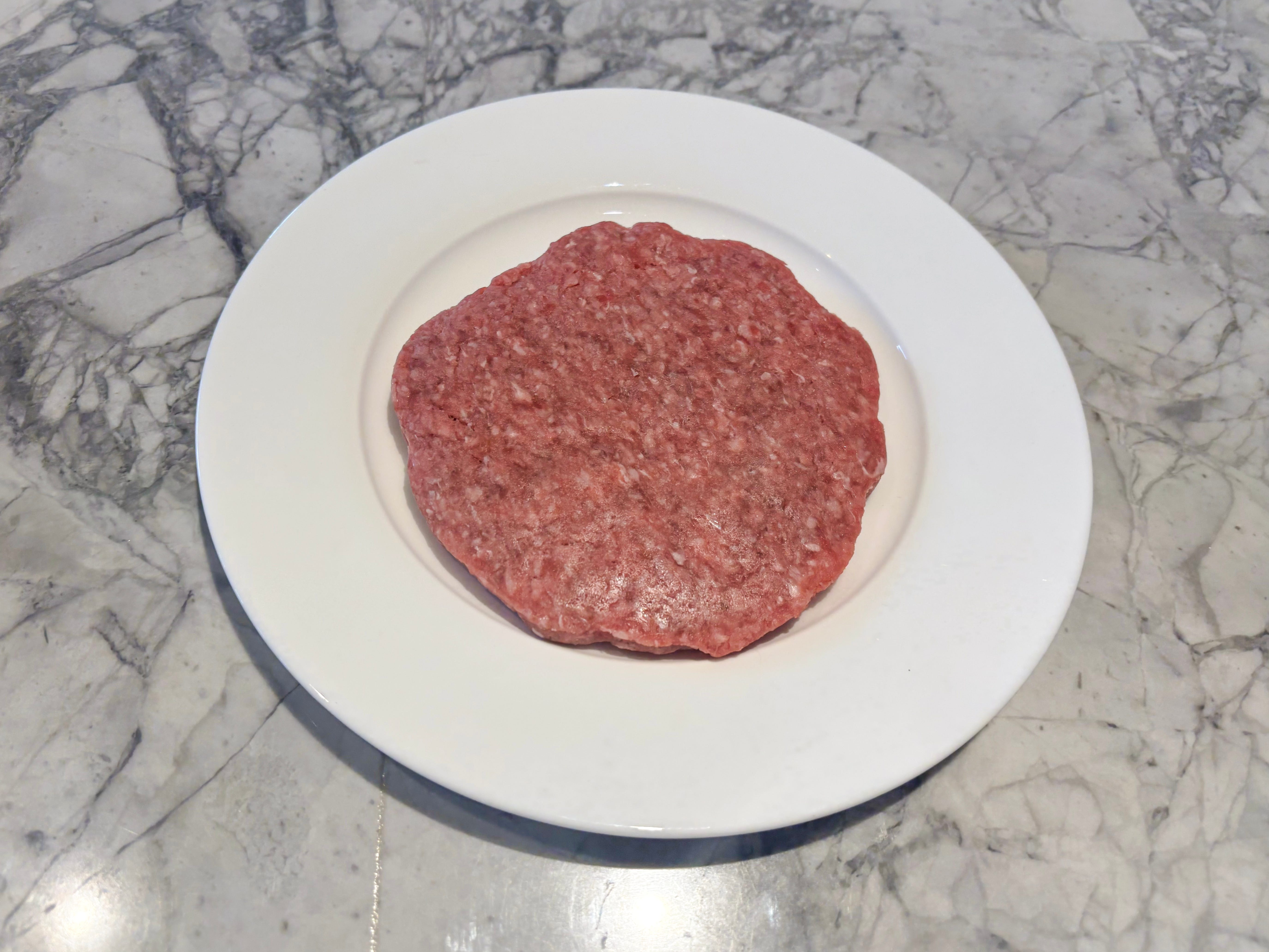 A pink raw beef patty on a white plate placed on a gray table