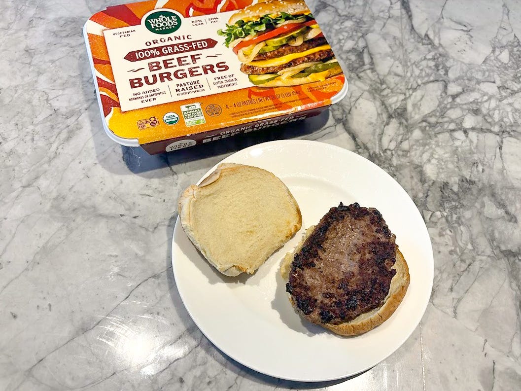 An orange box with a Whole Foods label and an image of a burger on it next to a white plate with a burger patty on a bun and the top of the bun placed on the side