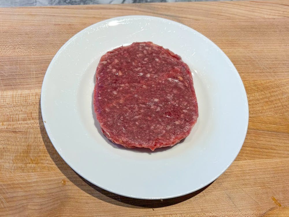 A round bright-red raw beef patty on a white plate placed on a wooden cutting board