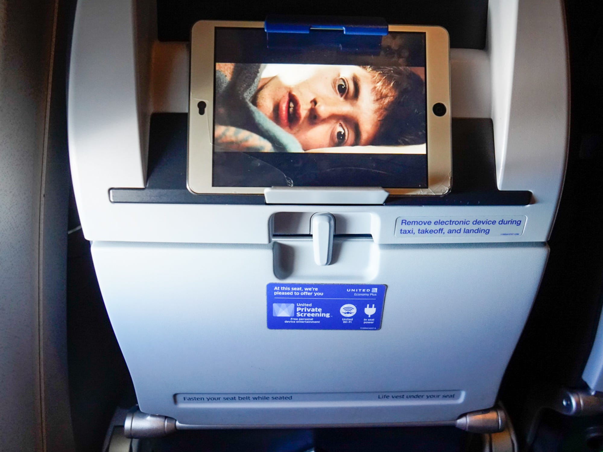An airline seat back holds an iPad playing a movie