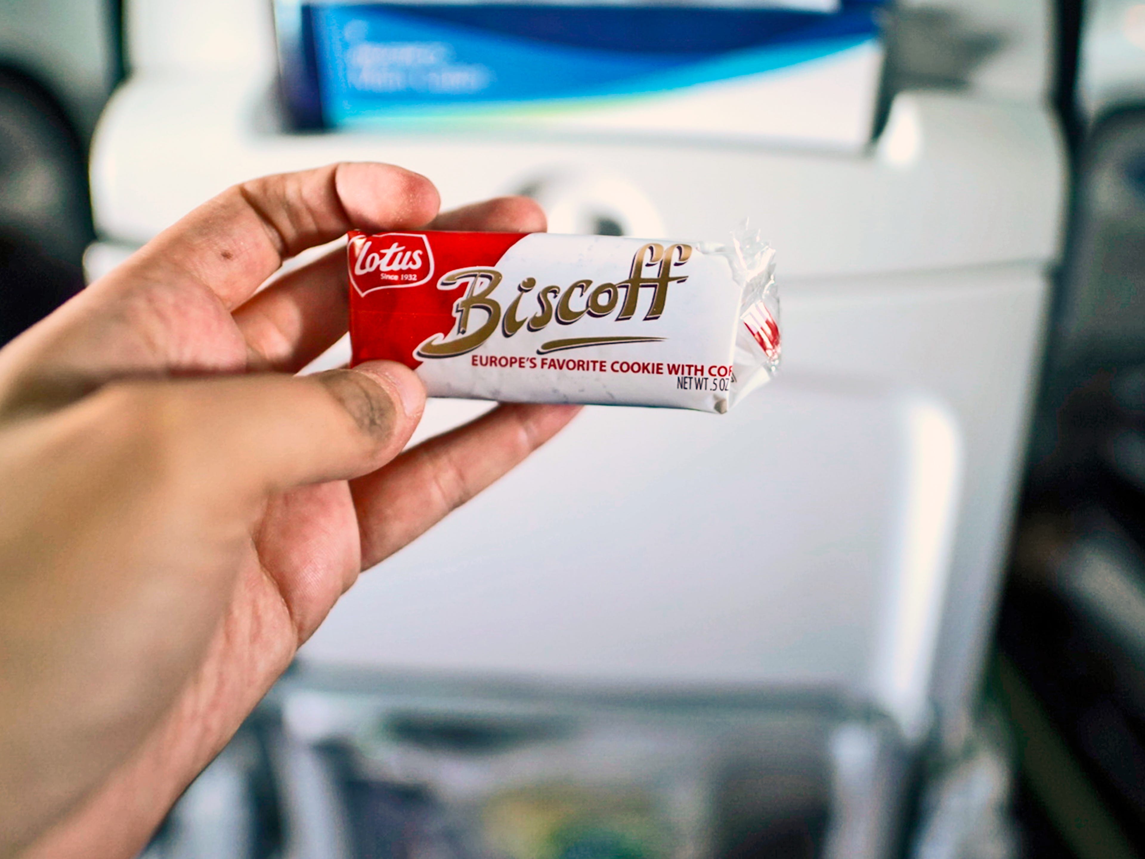 A hand holds a wrapped Biscoff cookie in front of a plane seat.