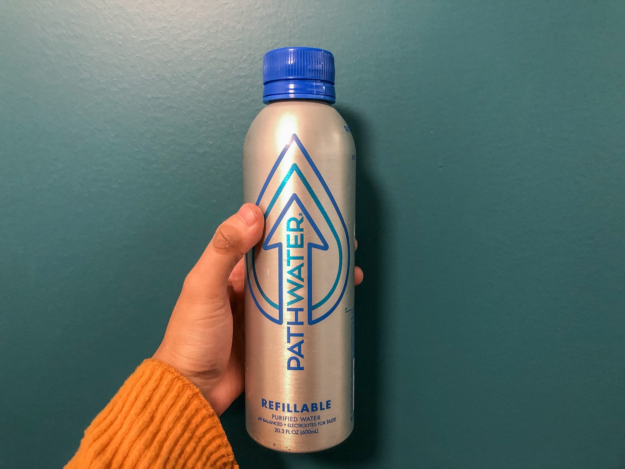 A hand holds a Pathwater refillable water bottle in front of a teal wall