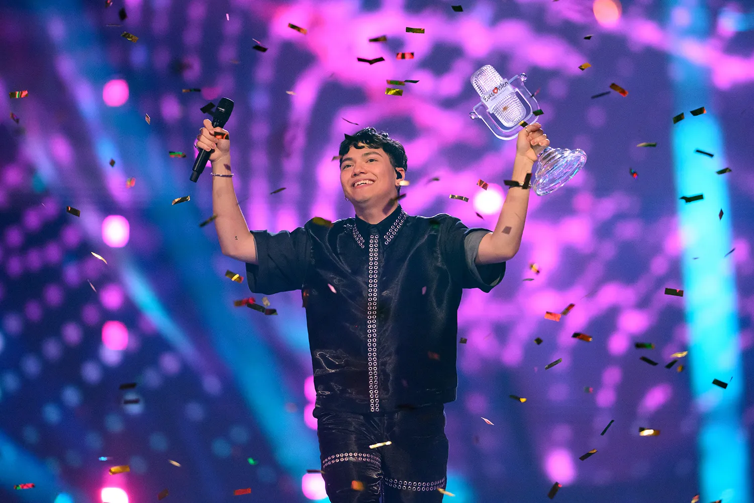 JJ holds up both hands one with a mic and one with a trophy as confetti falls on a stage.