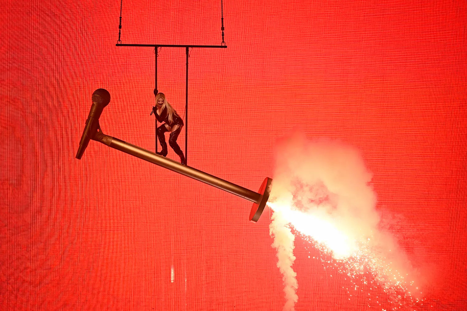 A woman performs atop a giant microphone shooting sparks.