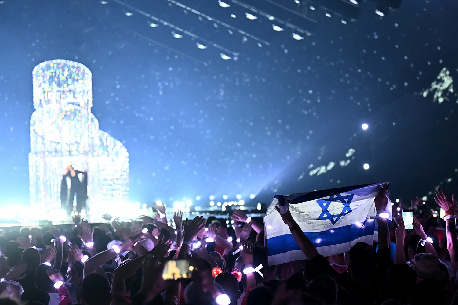 An Israeli flag is held aloft amid a large crowd with a singer on stage in the distance.