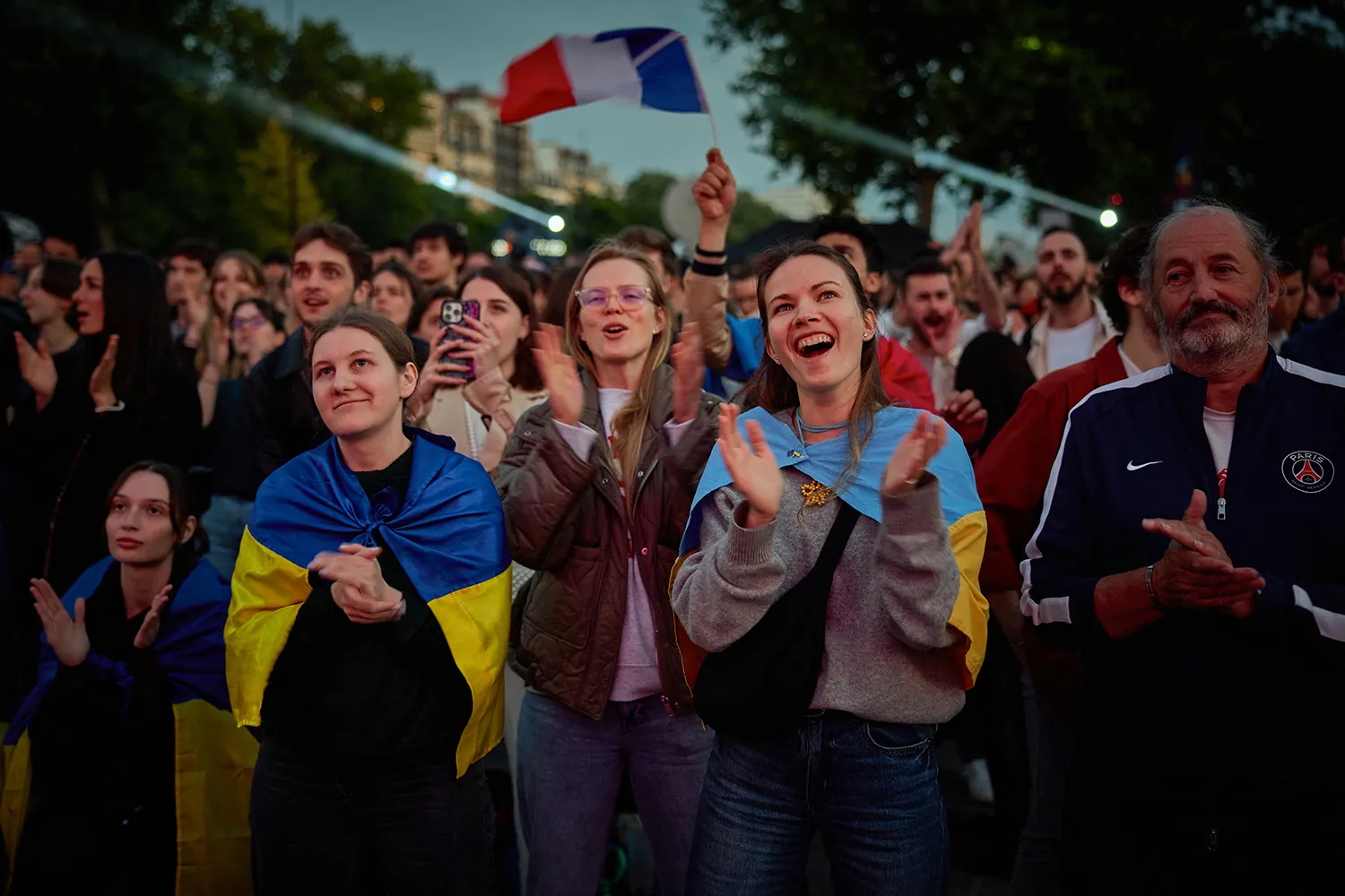 Fans draped in flags clap as they look up at a screen from a amid a large outdoor crowd.