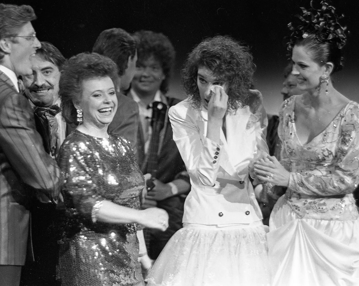 Celine Dion dabs her eyes with a tissue as people smile and embrace her on stage.