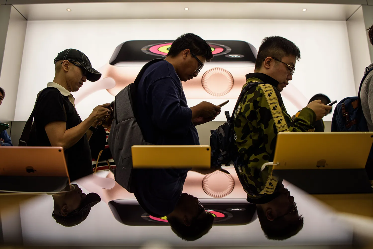 Three people in a line look down at their phones as they stand in front of a display with the latest iPhone behind them.