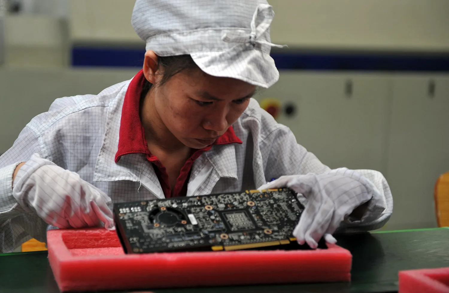 A worker wearing a white hat and white gloves leas in to look at a motherboard.