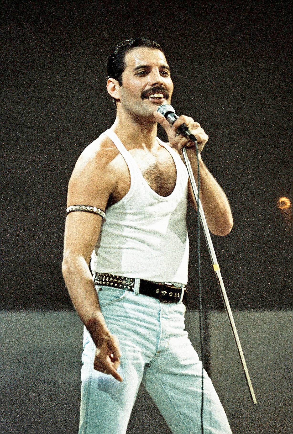 Freddie Mercury sings onstage in a white tank top. 