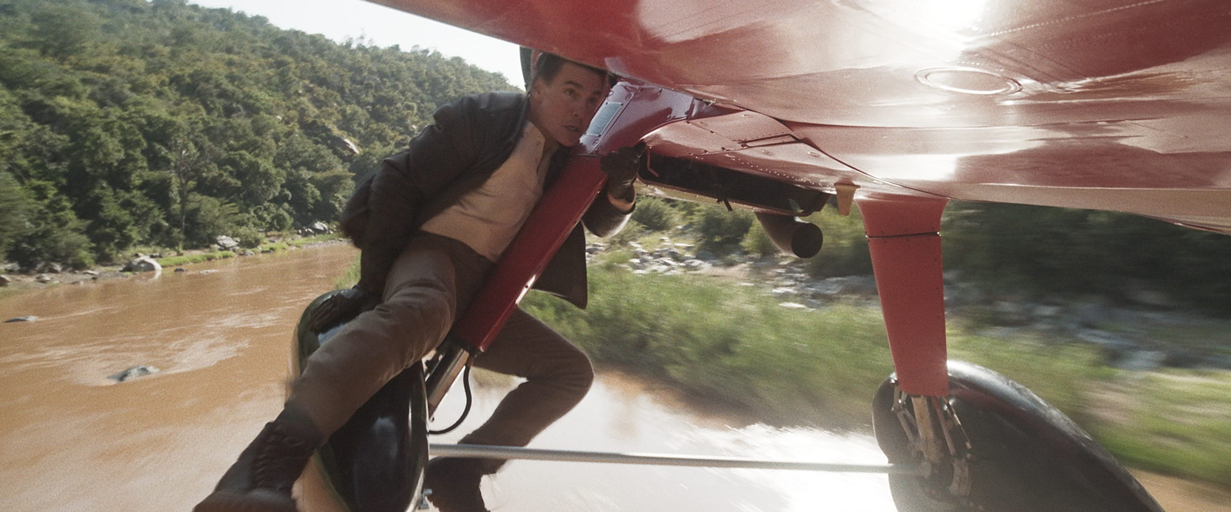 A man in a brown leather jacket and a white t-shirt holding onto the underbelly of a plane.