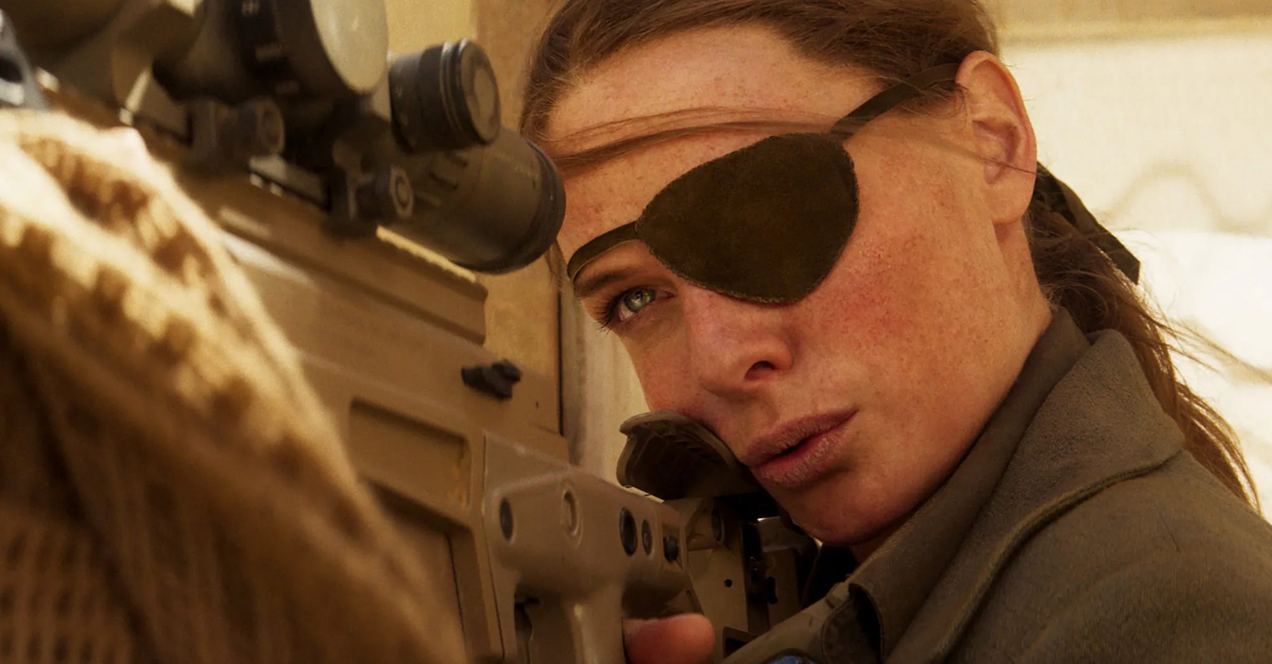 A woman with tied-back brown hair is wearing an eye patch and looking down the scope of a sniper rifle.