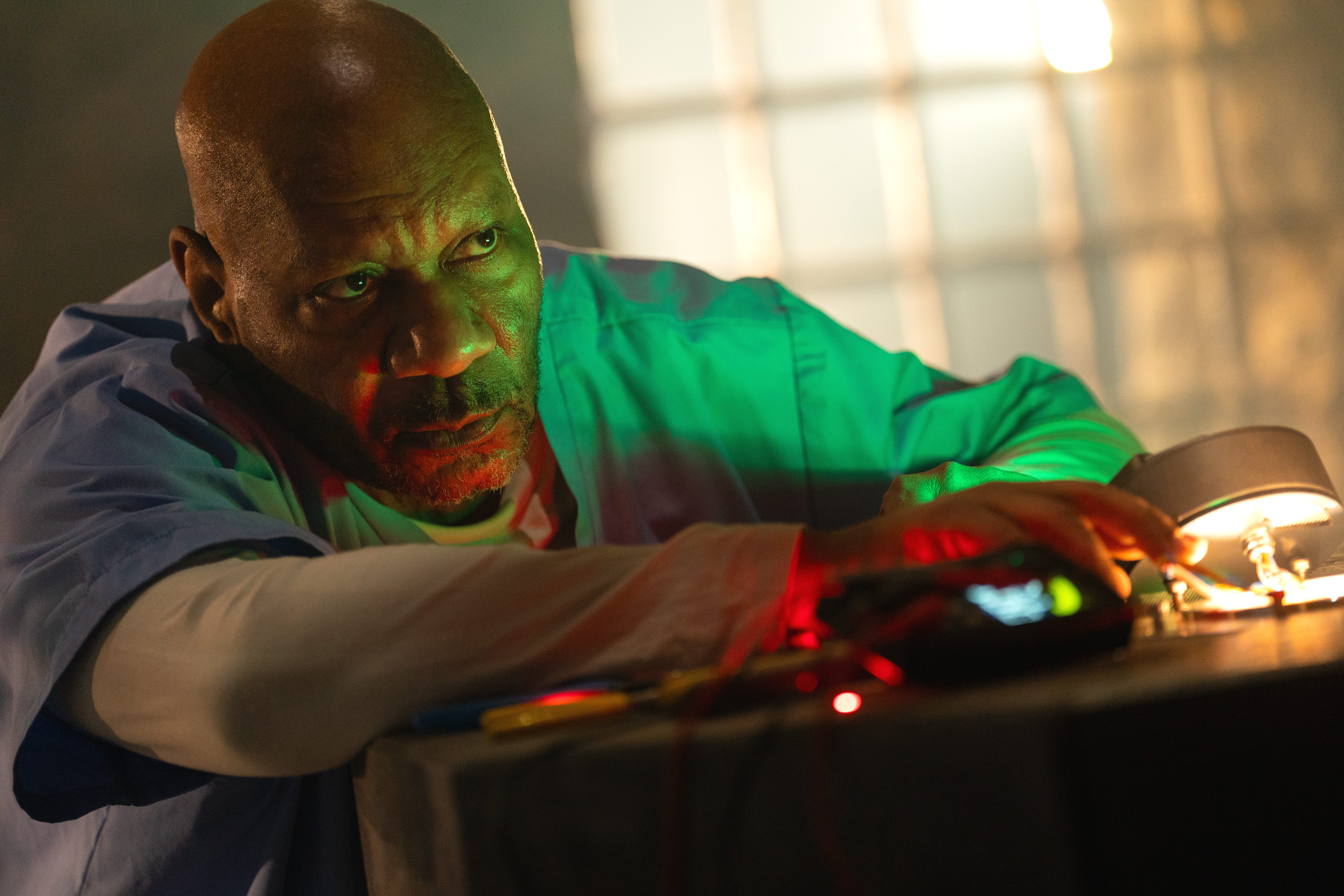 A bald Black man wearing blue medical scrubs while holding onto technical equipment on a desk.