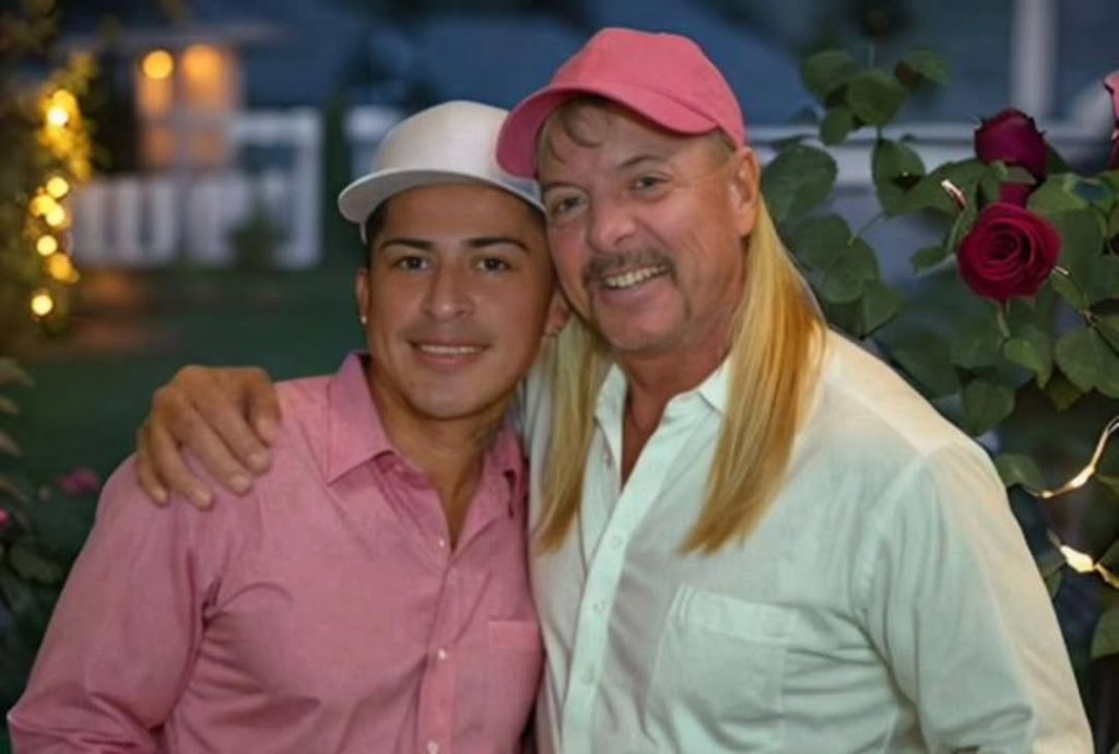 Jorge Flores Maldonado in a pink button down shirt and white baseball cap and Joe Exotic with a white button down and pink baseball cap and his arm around maldanado posing together and smiling at the camera with roses and lights behind them