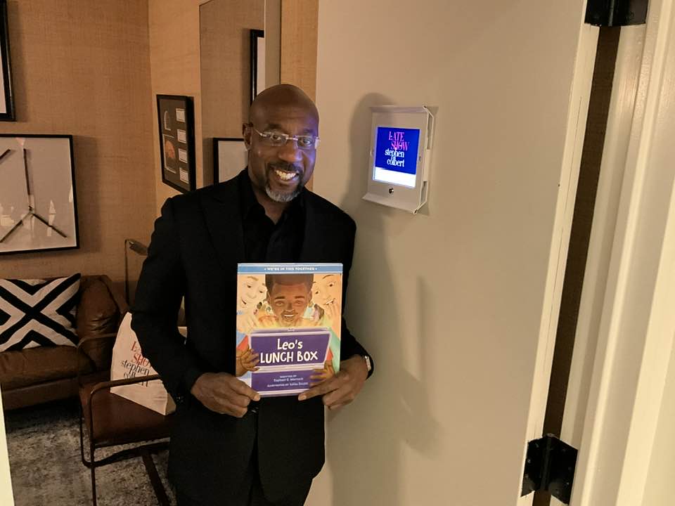 U.S. Senator Raphael Warnock smiling holding a copy of his book 'Leo's Lunchbox' while looking at the camera