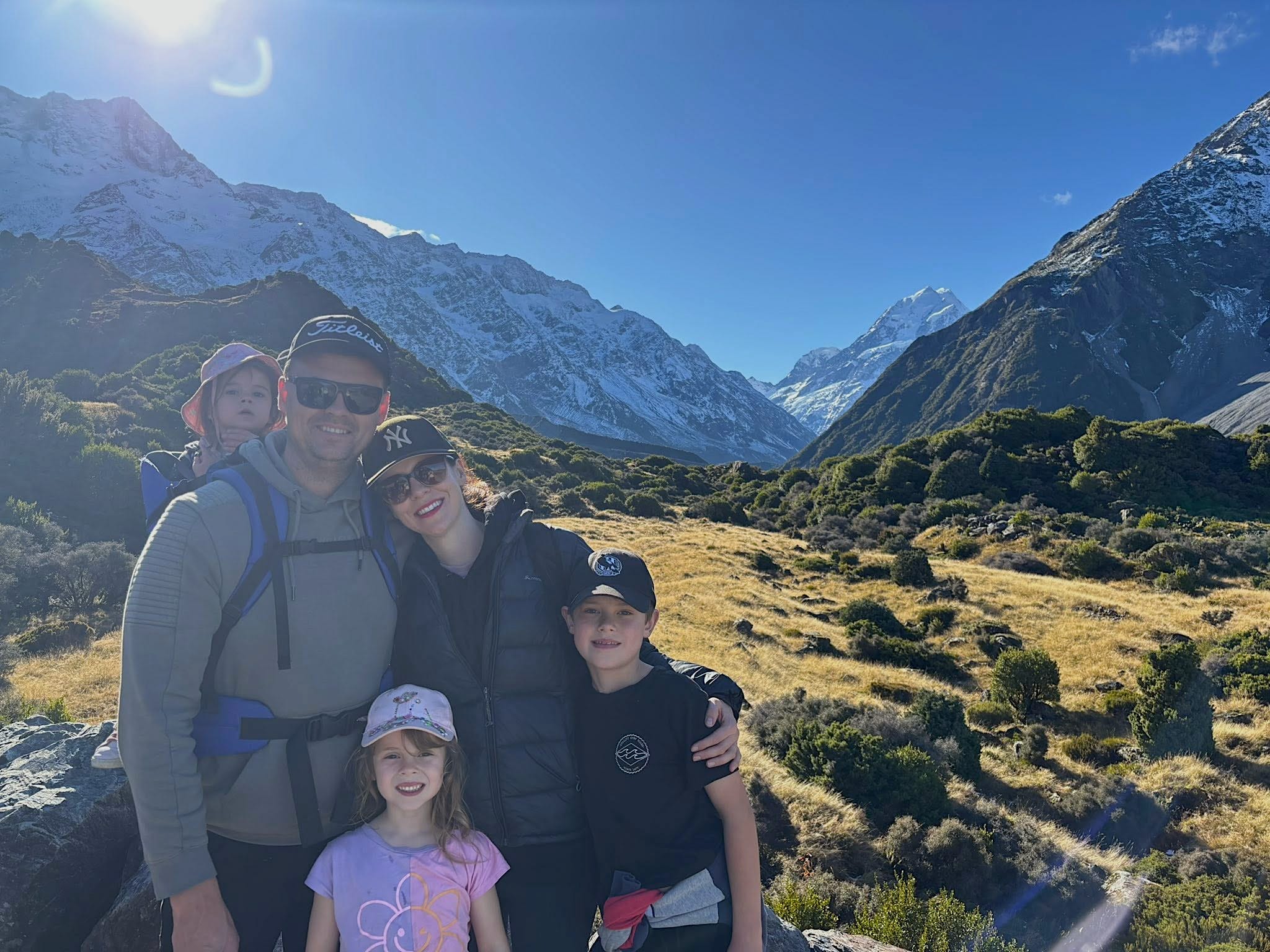 The author and her family hiking Mount Cook while on their trip in New Zealand. Grass and a blue sky and mountains are behind the family of five.