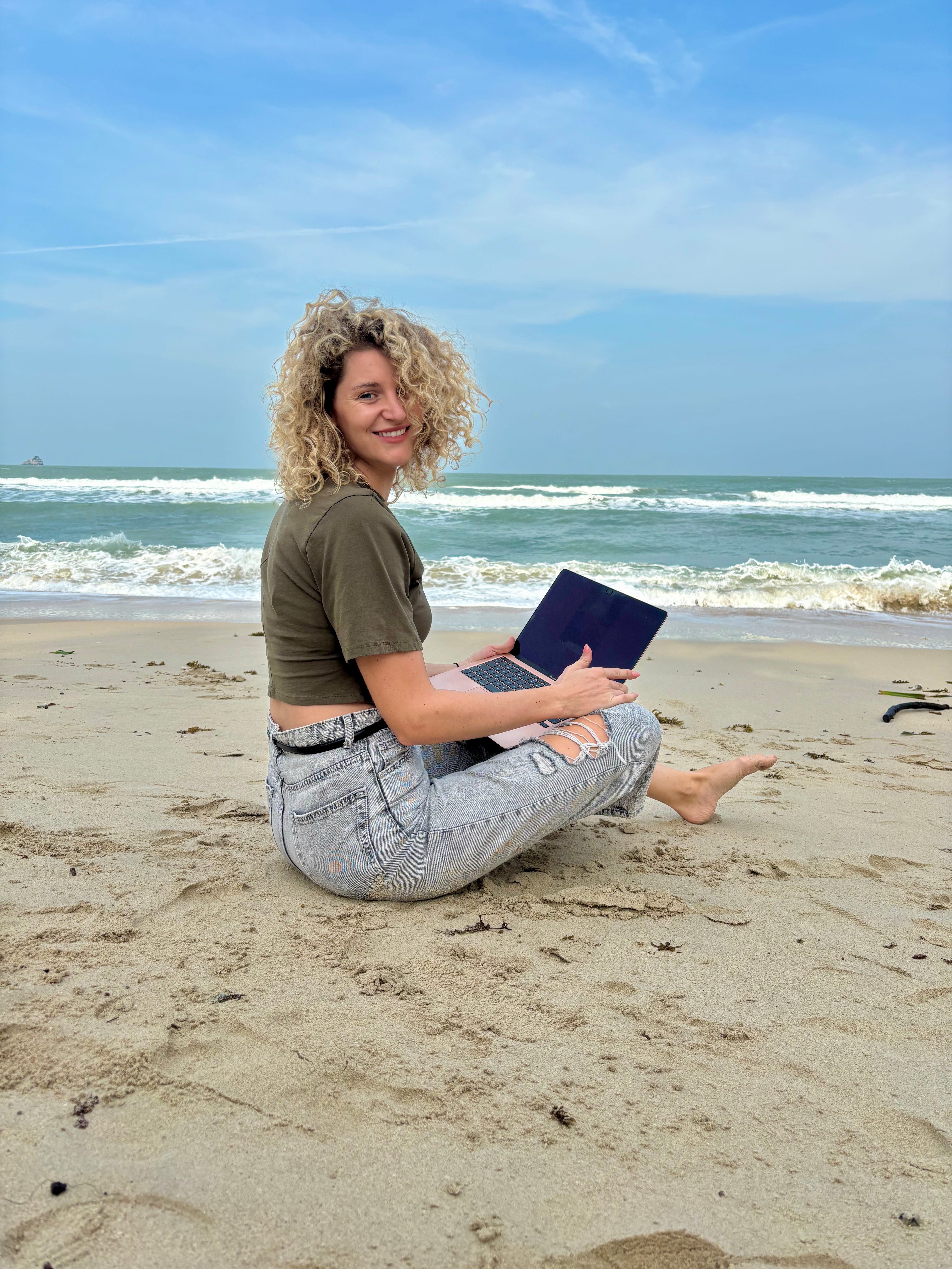 A woman sitting on the sand, by the ocean, in Koh Samui, Thailand, with her laptop.