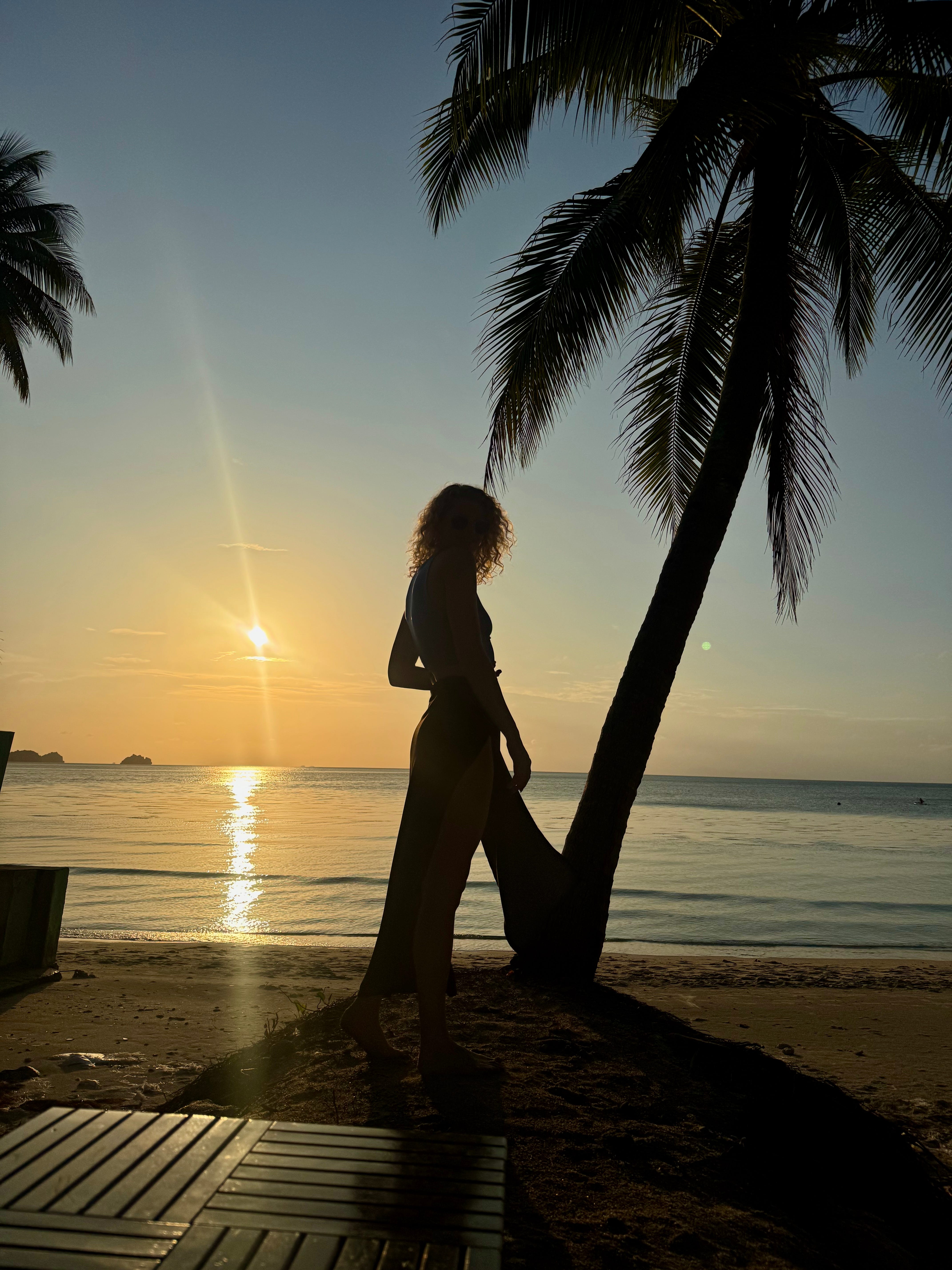 The silhouette of a woman standing by a tree, watching the sunset at a beach in Koh Samui, Thailand.