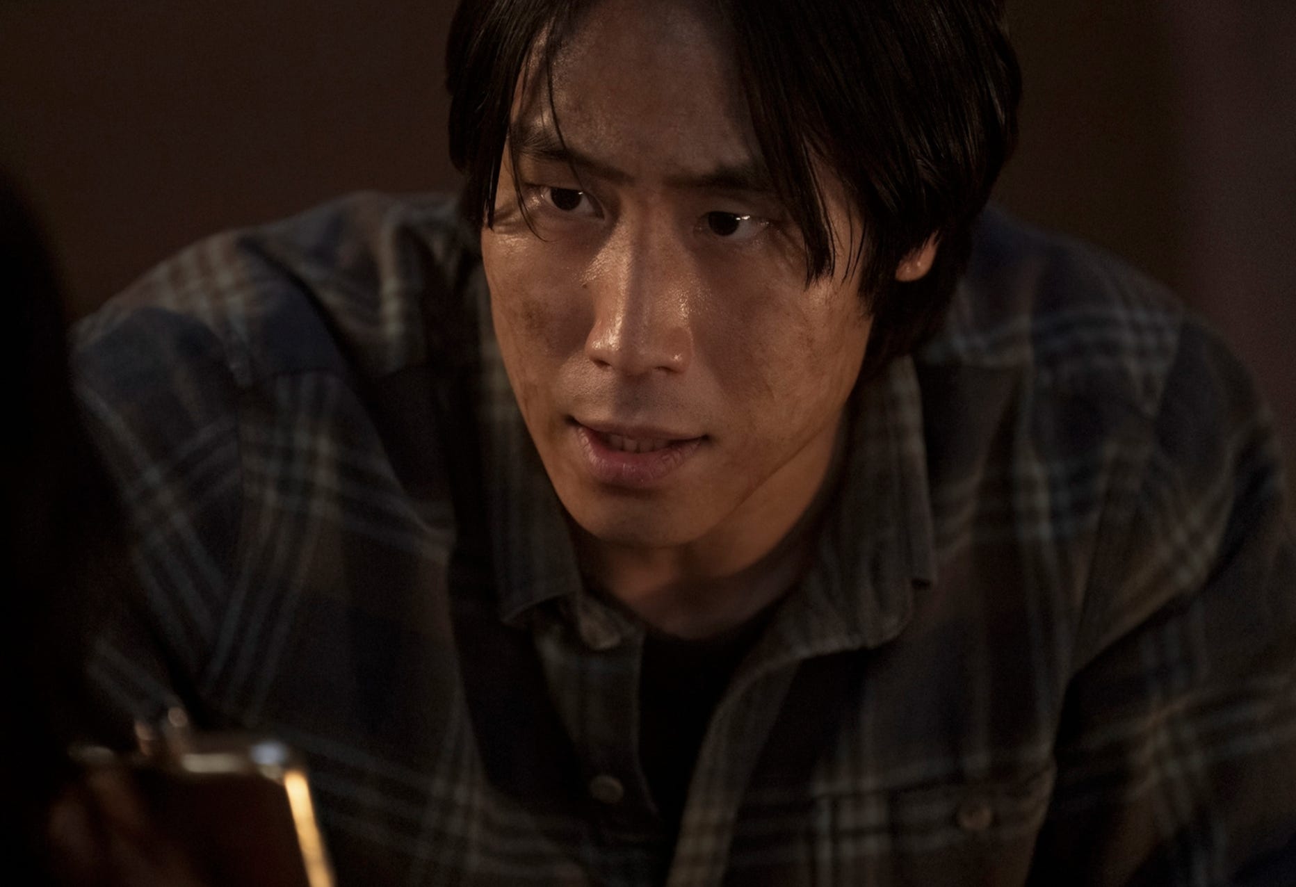 An Asian man with medium-length black hair is wearing a blue and brown plaid shirt and is holding a silver hipflask, his face is dirty.