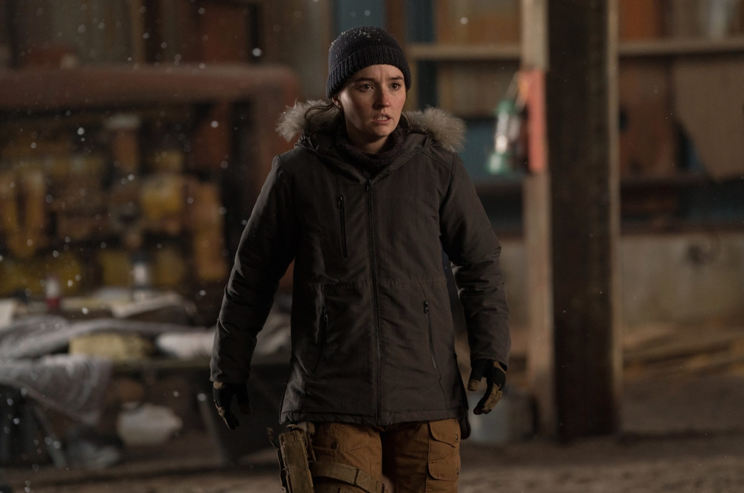 A young woman dressed in a black parka and wearing brown tactical trousers with a gun strapped to her left leg. She's wearing a black wooly hat. She's stood inside a warehouse.