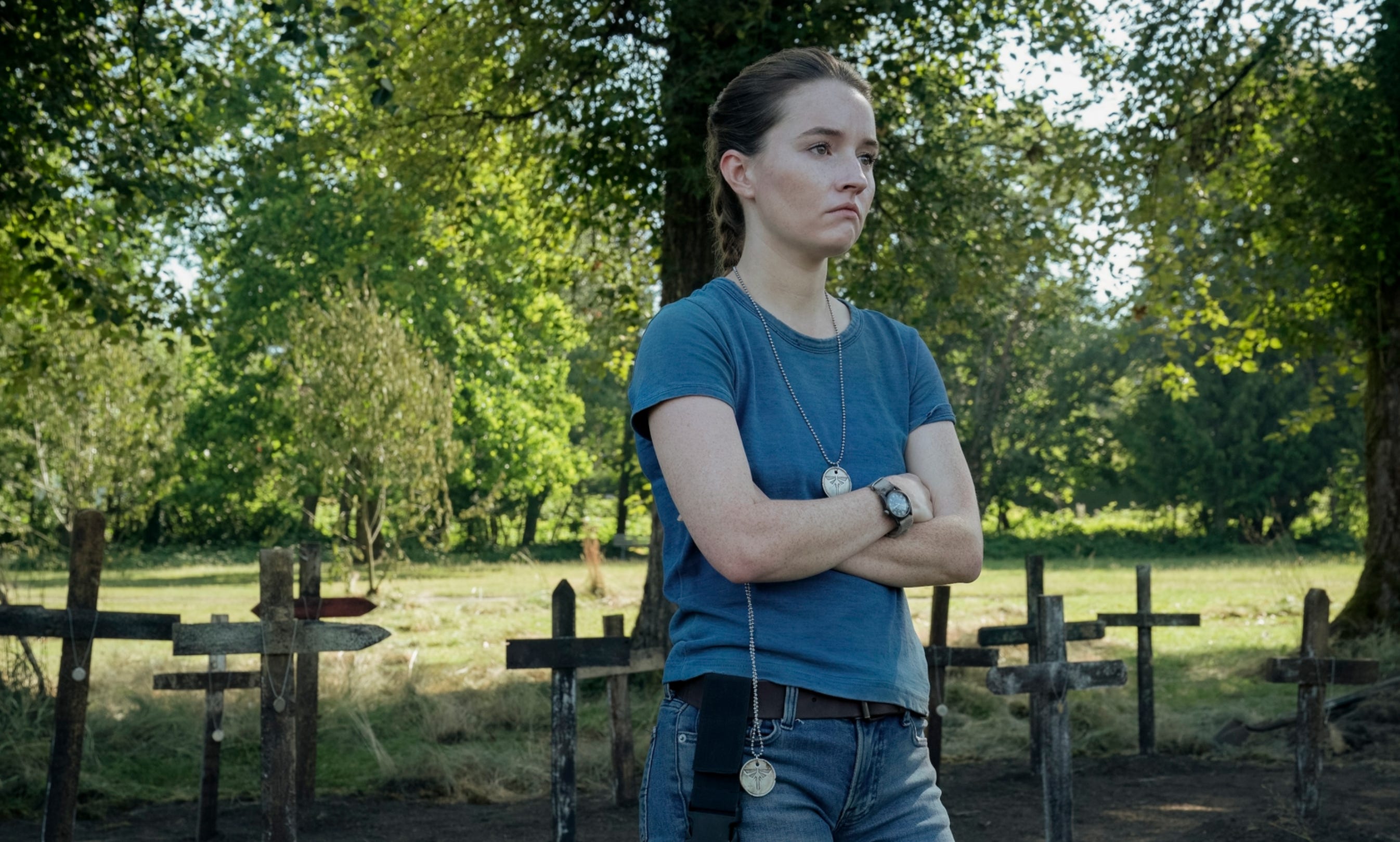 A young woman with brown hair tied back in a ponytail. She's wearing a plain blue t-shirt, blue jeans and a brown belt. There is a green and black watch on her left wrist. There is a silver medallion hanging around her neck. There are wooden crossed on the ground directly behind her, with a field that has several trees in it further in the background.