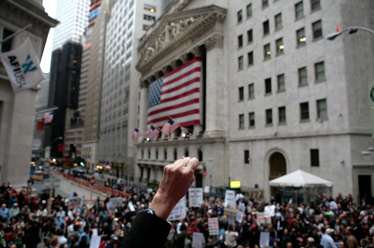 A person raises a fist in the foreground as a crowd gathers outside the New York Stock Exchange on Wall Street, where a flag hangs on the front of a building.