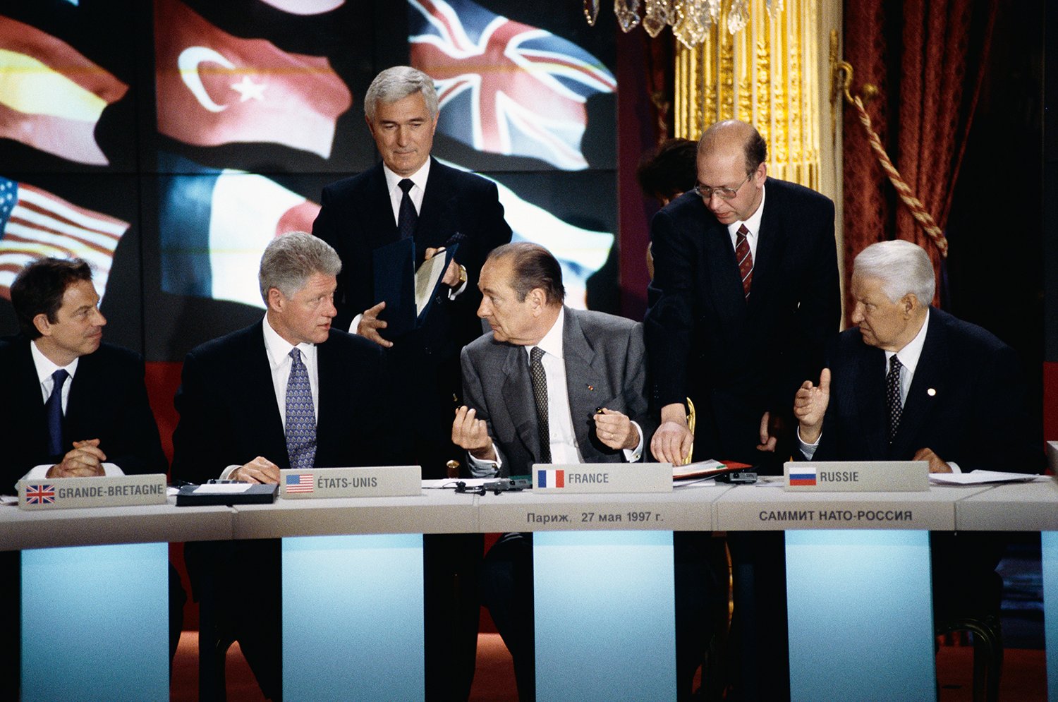 Two men sit at a table as two others stand behind them. Flags of various countries are on a sign or screen behind them.