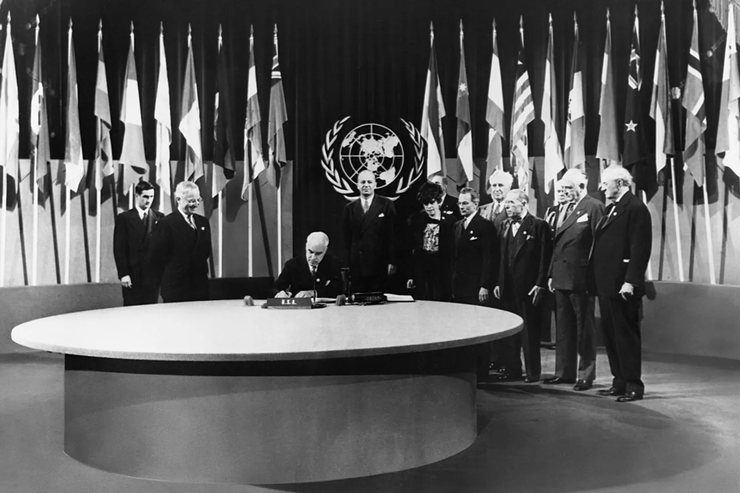 A man sits at a large round table as other men stand on either side of him. The seal of the United Nations is behind them flanked by many country flags on stands.