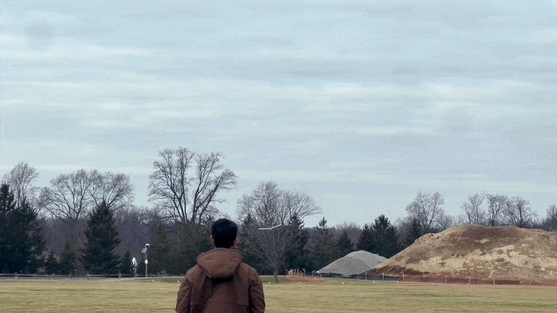 gif shows drone with fixed wings and rotors flying above a field in front of a young man in a brown jacket