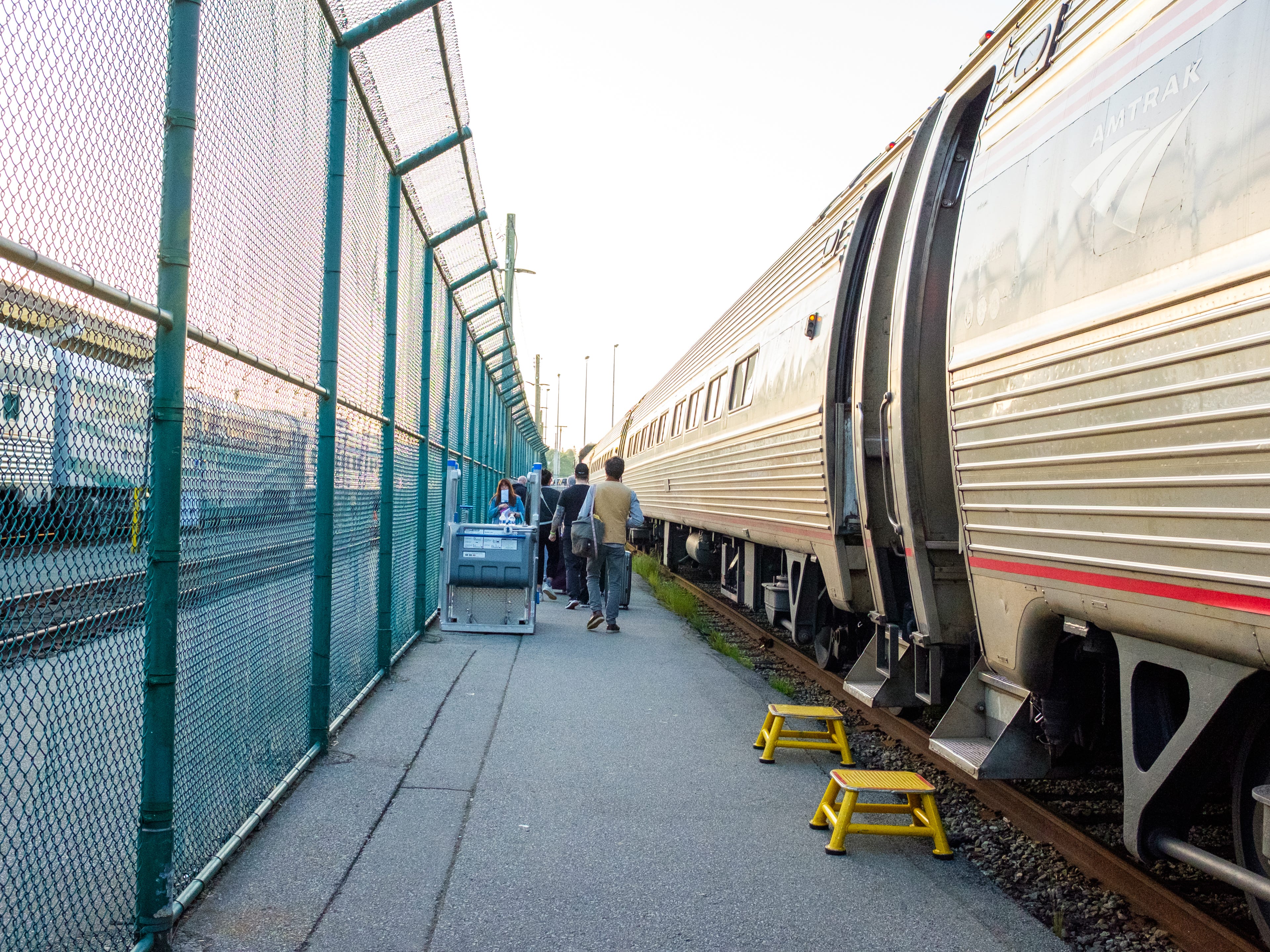 Passengers walk down a train platform with a train on the right and a fence on the left