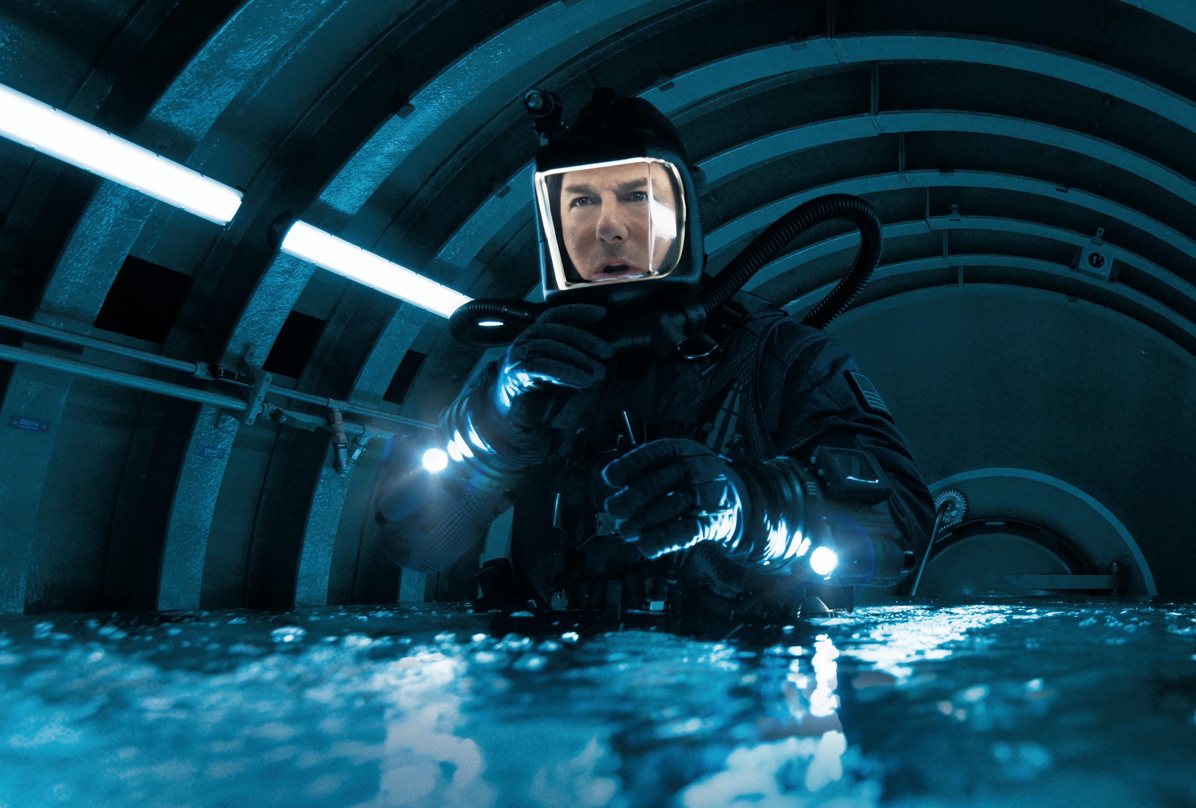 A man in a scuba diving outfit with an illuminated mask is stood in a circular room that is filling up with water.