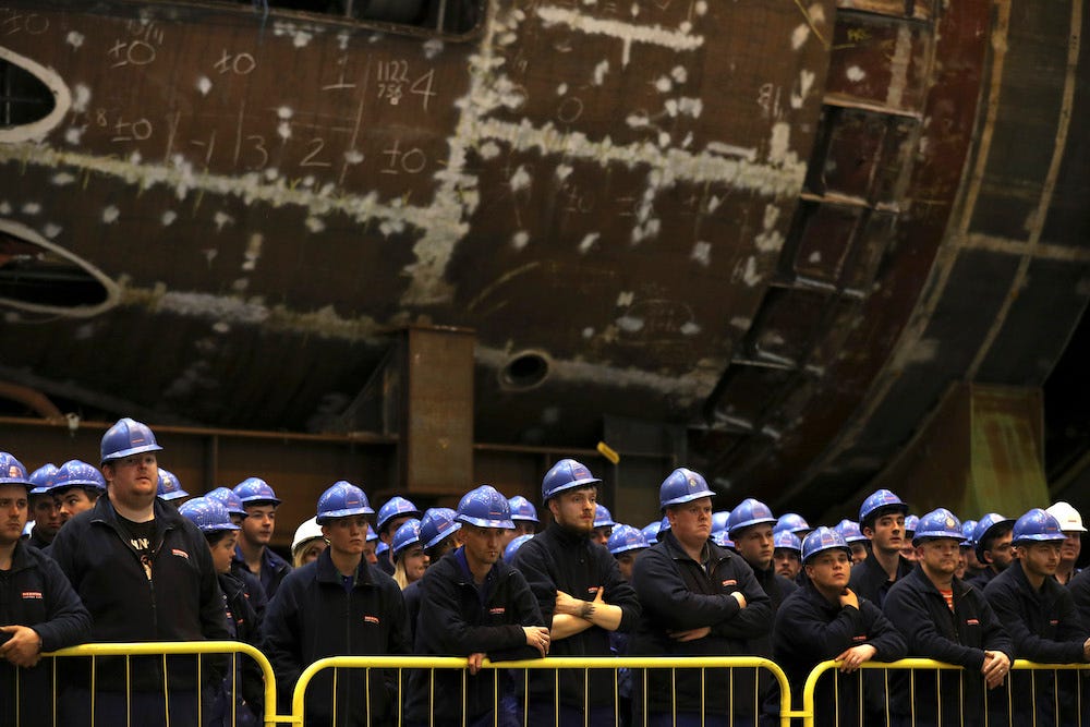 Workers listen, with a large metal keel behind them, as Britain's Defence Secretary Michael Fallon speaks after watching the first piece of steel for the successor submarine programme being cut at BAE Systems on October 5, 2016 in Barrow-In-Furness, United Kingdom.
