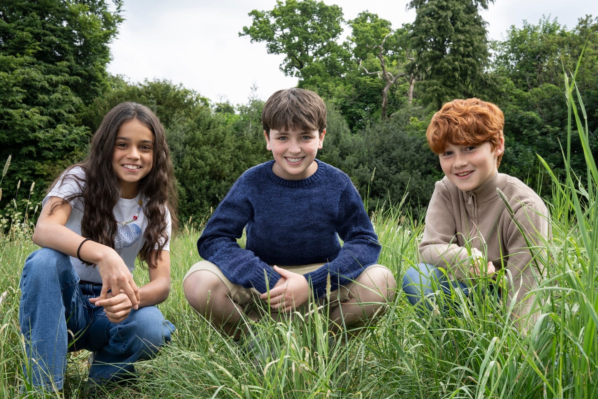 A young girl and two young boys crouching in a field and smiling.