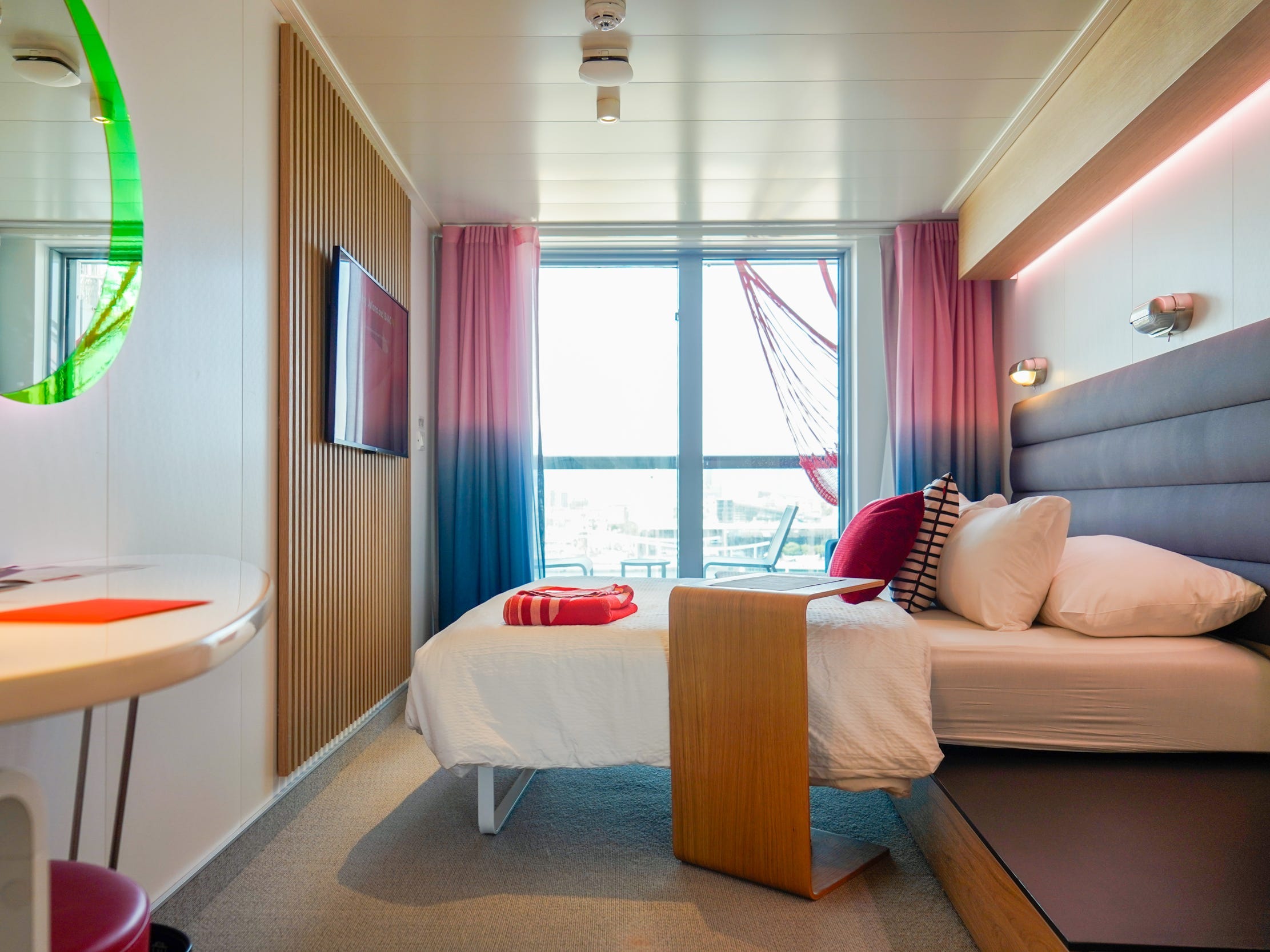 A bed with a navy headboard, white sheets, and white and red pillows as well as a red bolded blanket at the bottom seen from the side in front of a large window with red and blue curtains. There's a mountain TV on a wooden-planed wall to the left  and a wooden side table next to the bed.