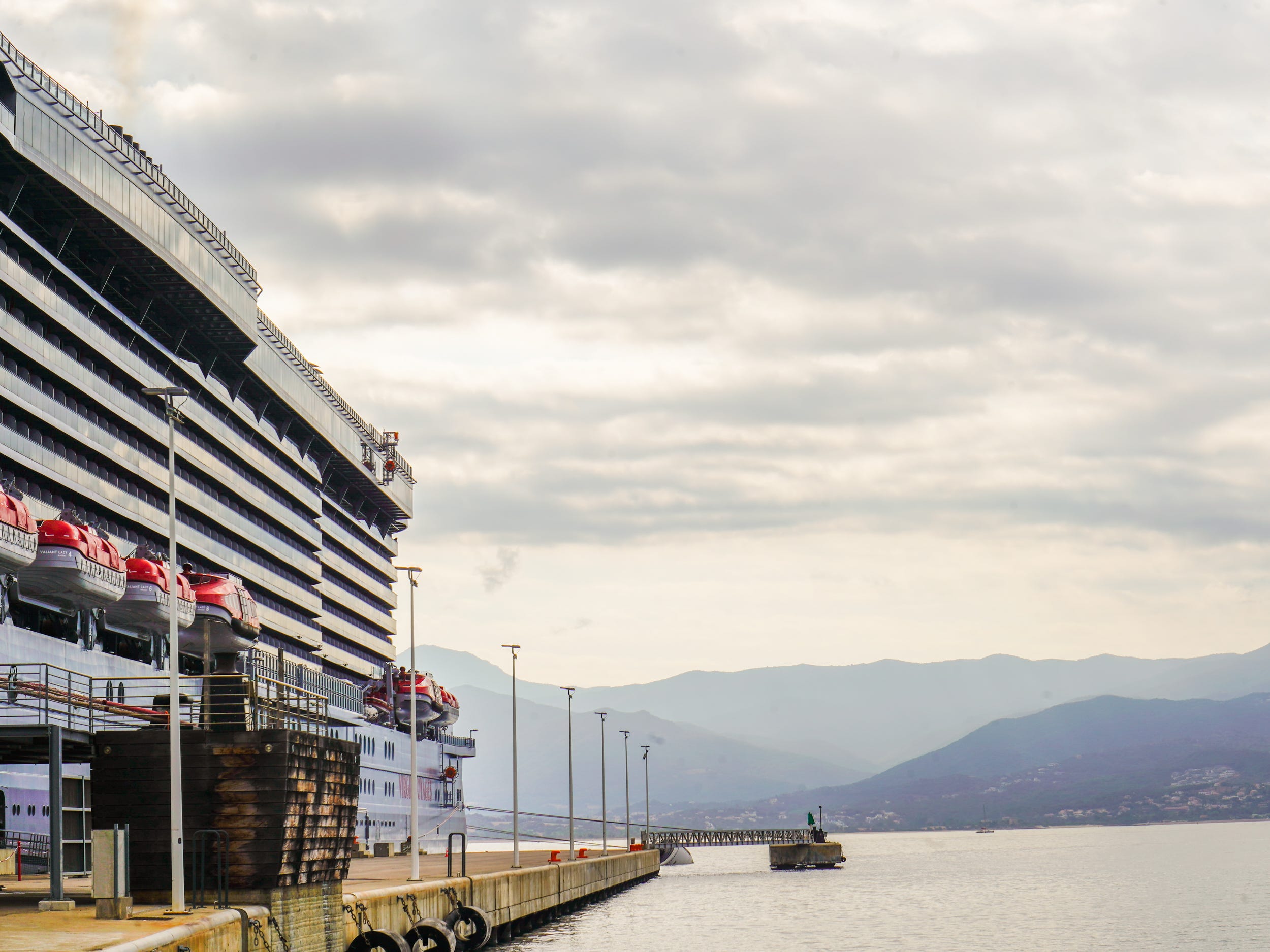 The end of a cruise ship come out of the left with the ocean on the right. There's a mountain range and cloudy skies in the background