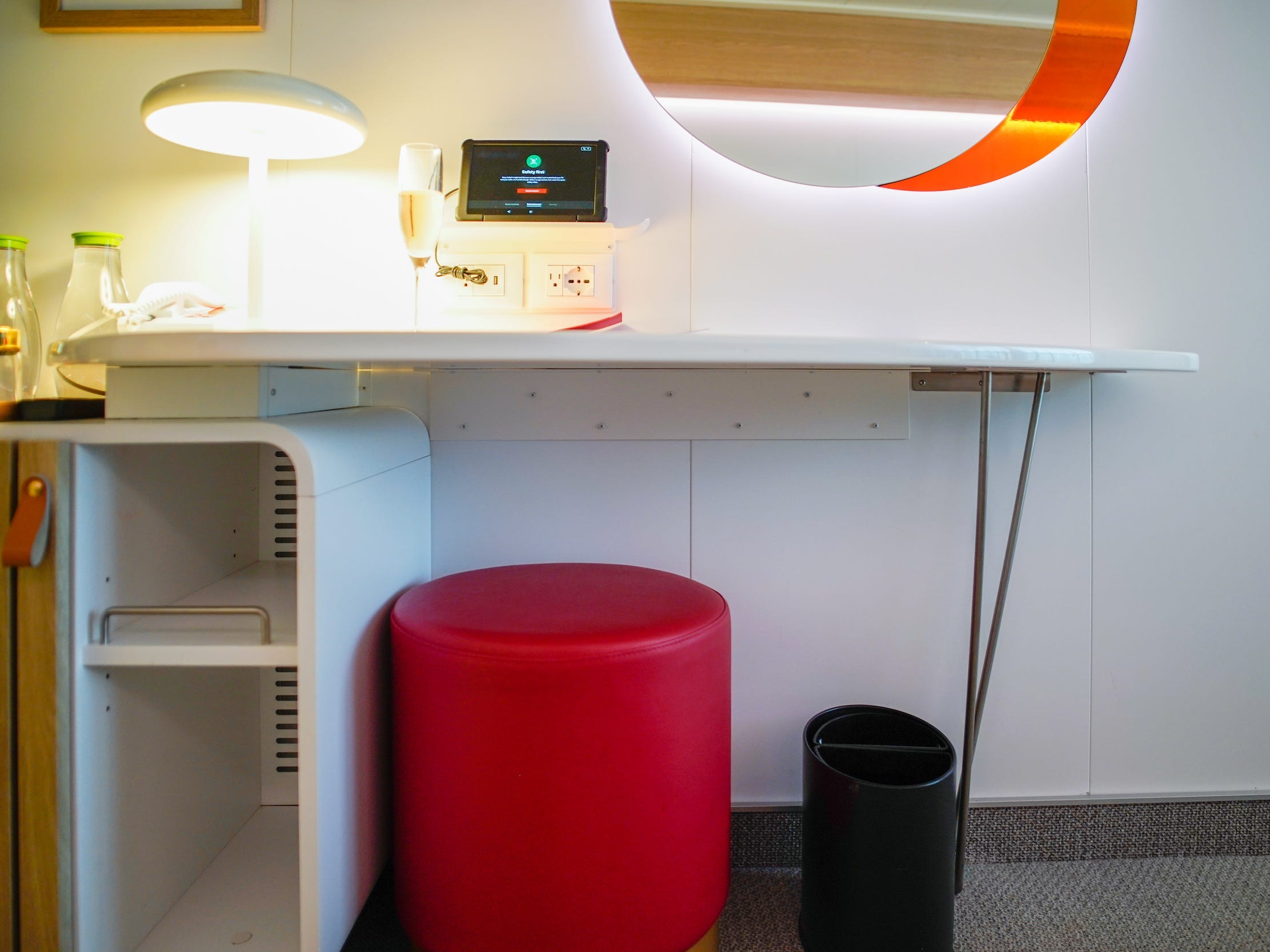 A white desk with a mirror above it and a lamp on top. There's a red cylinder-shaped ottoman below it