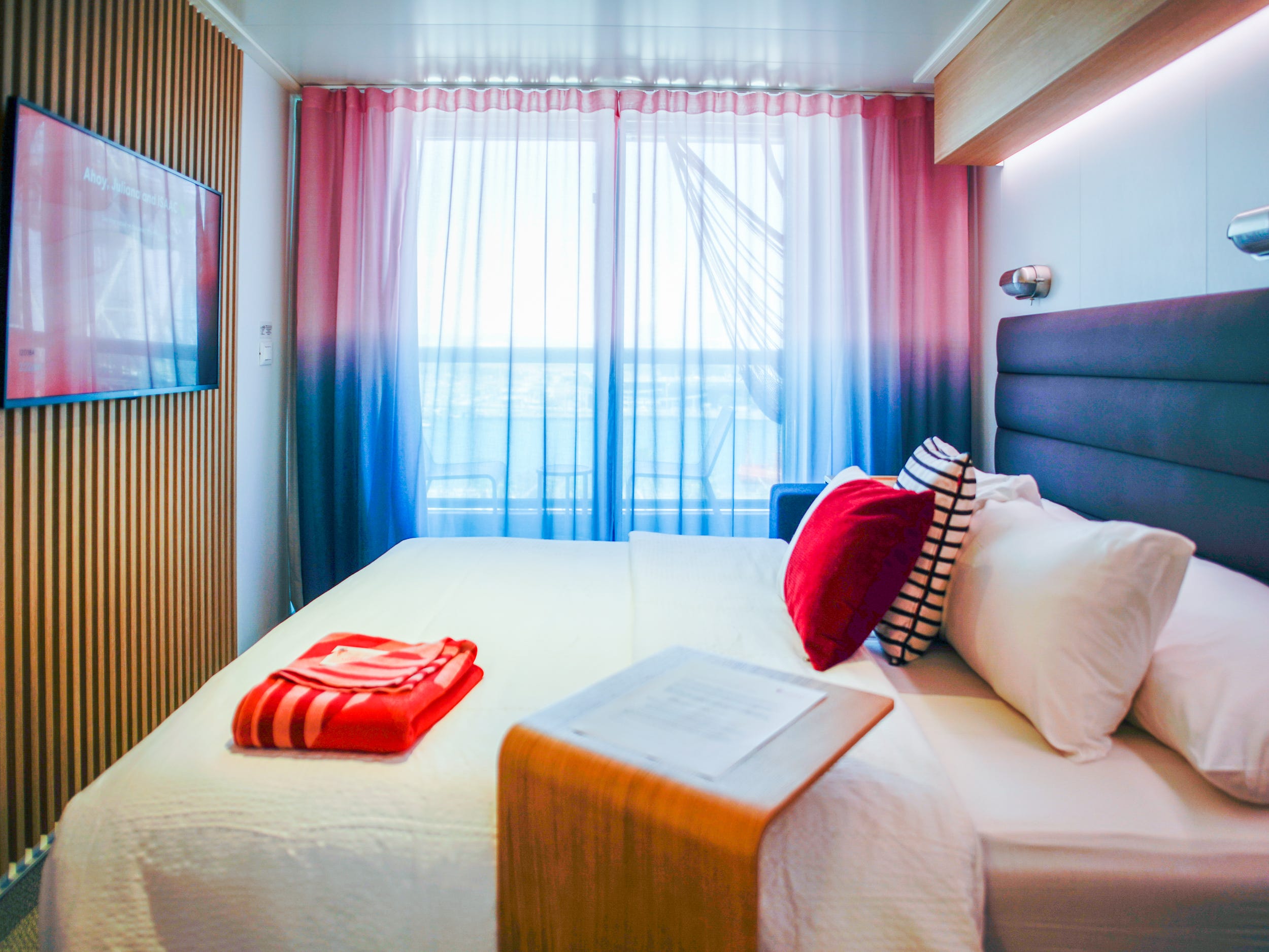 A bed with a navy headboard, white sheets, and white and red pillows as well as a red bolded blanket at the bottom seen from the side in front of a large window with red and blue curtains. There's a mountain TV on a wooden-planed wall to the left.