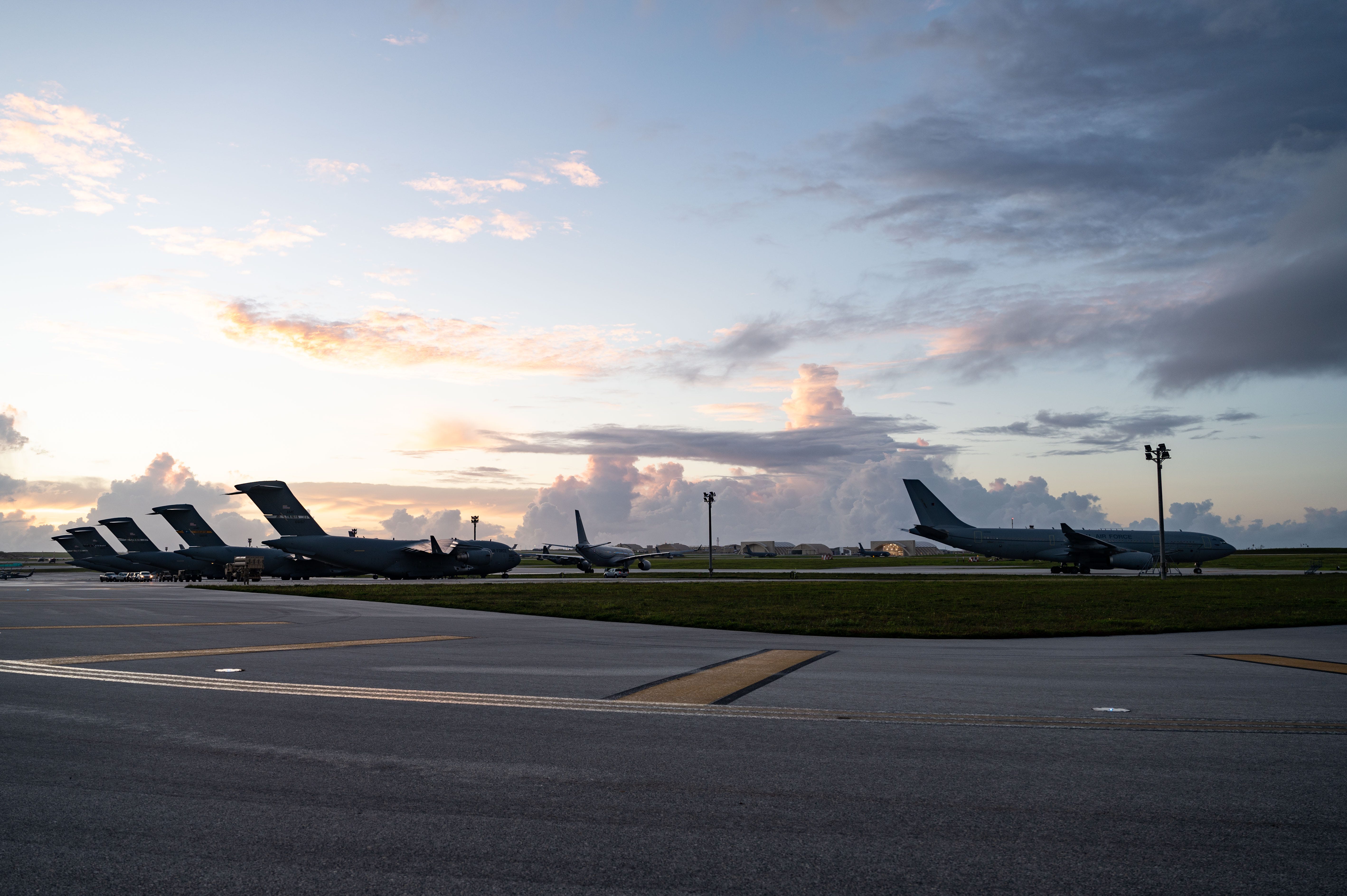 Various military aircraft are parked at an airbase below a sunset and some clouds.