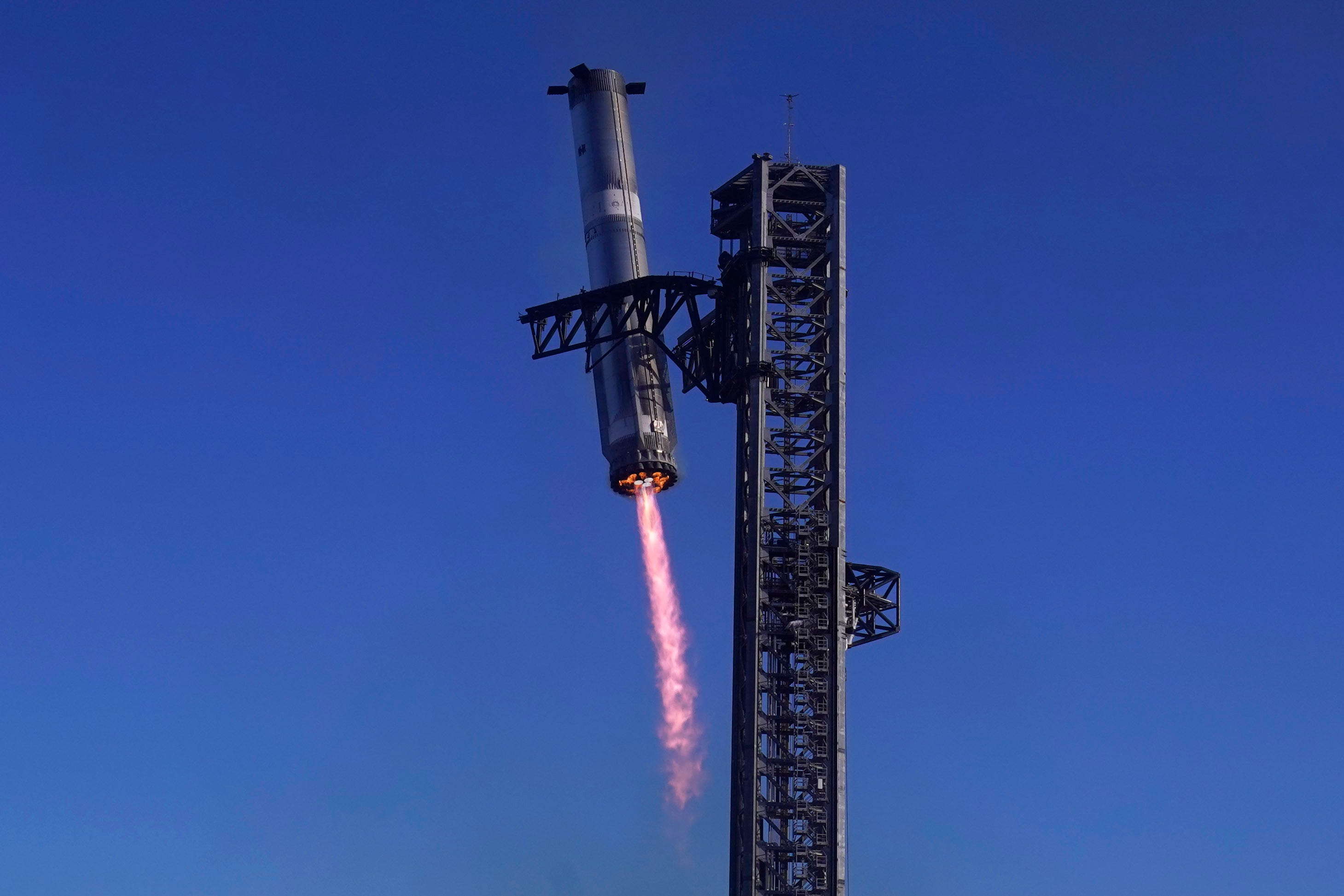 super heavy rocket booster long metal cylinder with bottom engines firing shooting out long red flare as the booster lowers itself into the metal arms of a launch tower