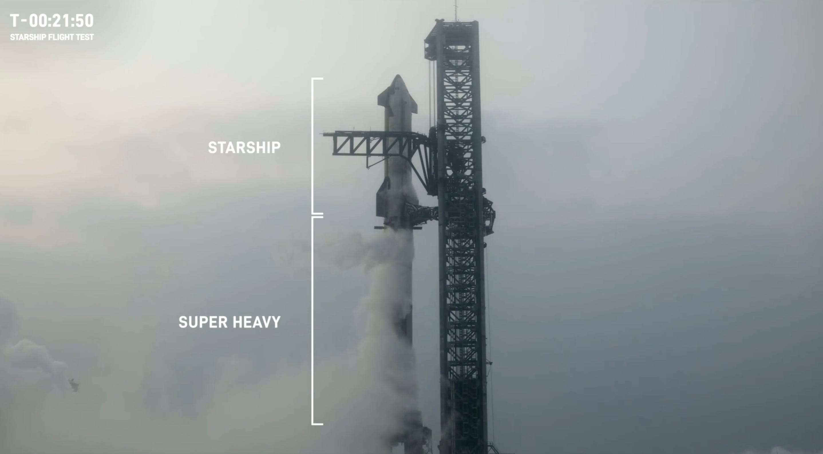 Starship atop its Super Heavy booster next to a tall launch tower in the foggy sky.