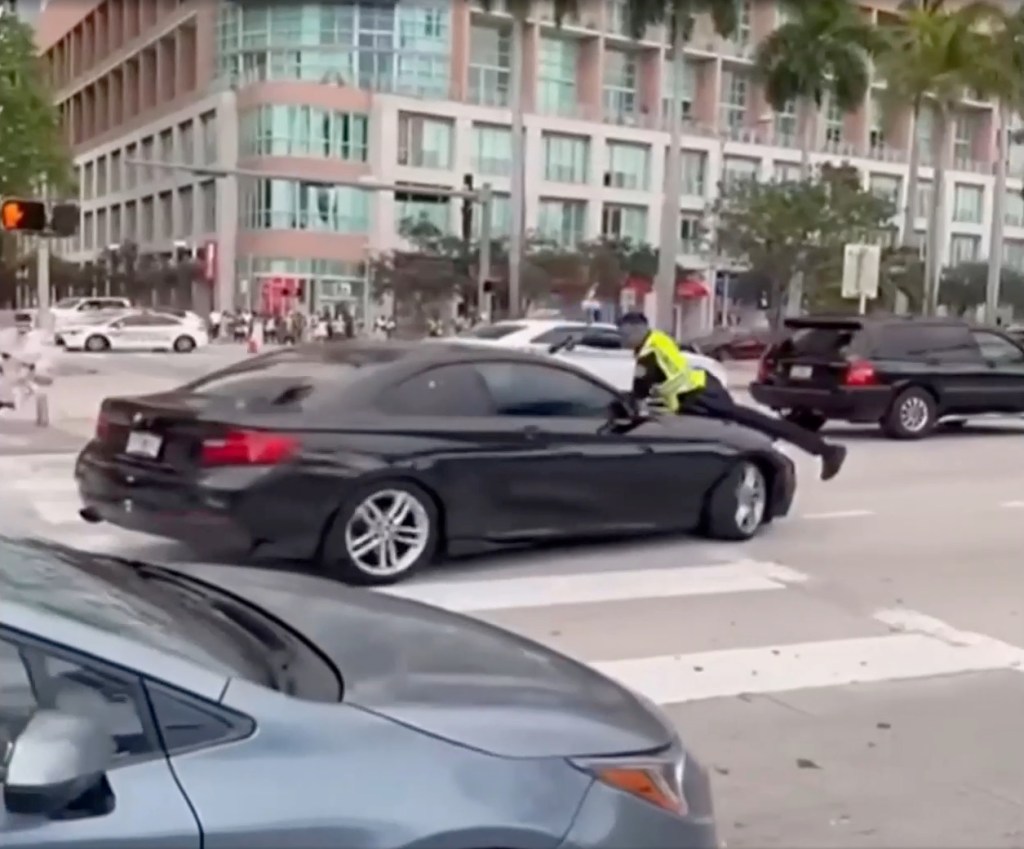 A Miami police officer was captured firing multiple rounds through the windshield of a car as he clung to its hood for dear life after the driver allegedly hit him and accelerated forward.