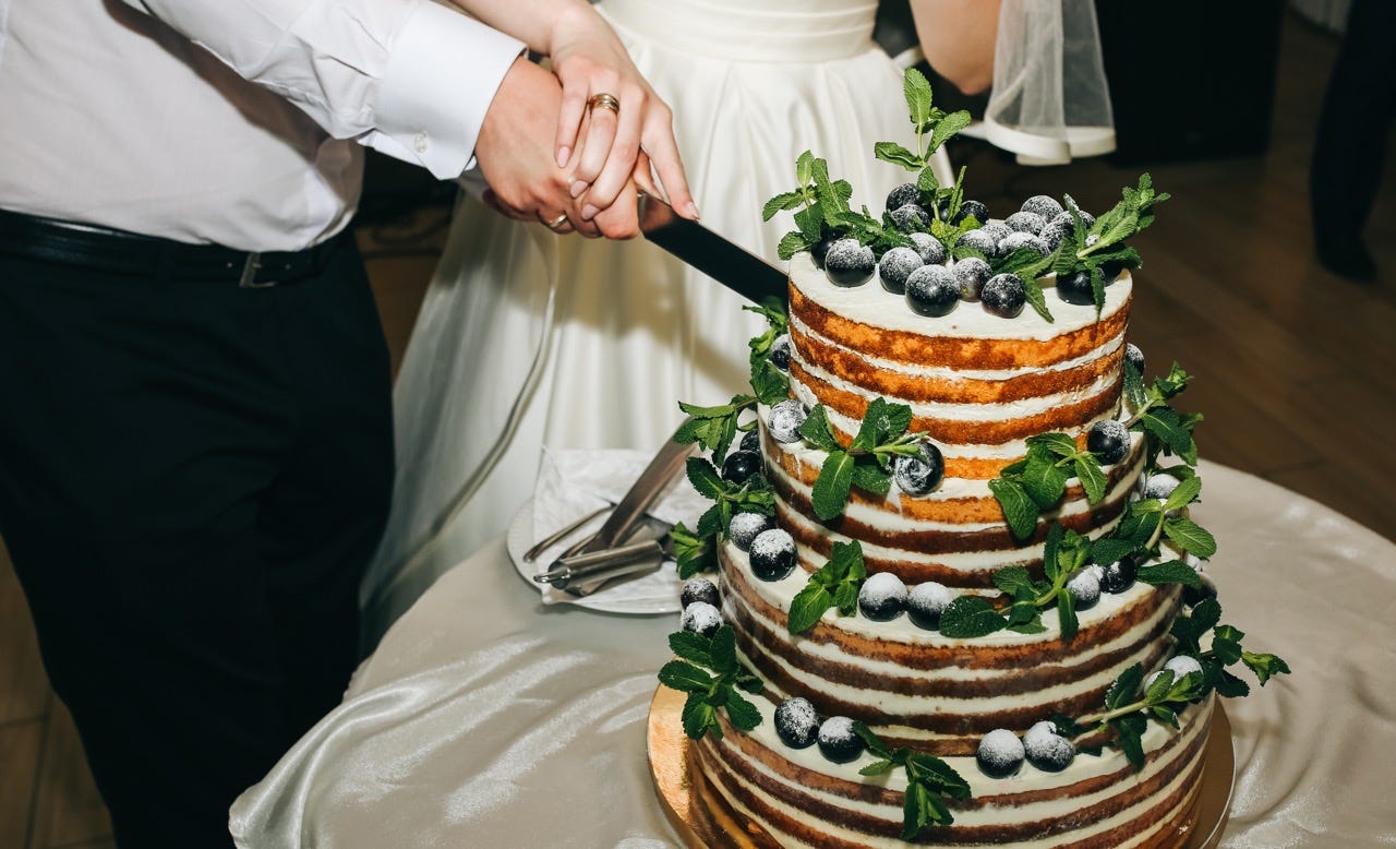 Couple holding knife together at wedding cutting a tiered cake with fruit and greenery on it