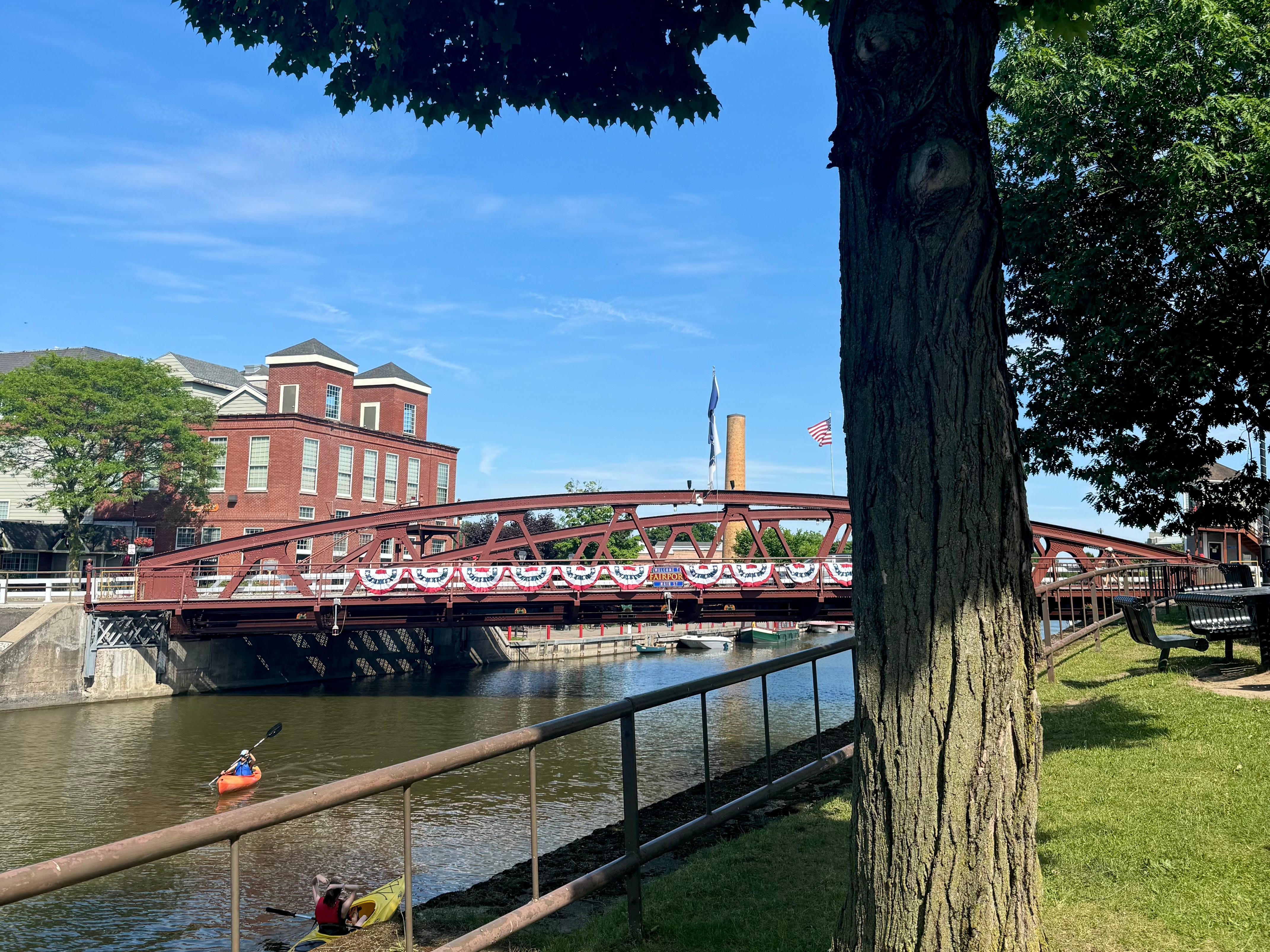 A wide shot of the Erie Canal with a large tree in the foreground. Kayakers are on the water and there's a bridge decorated with red, white, and blue banners.