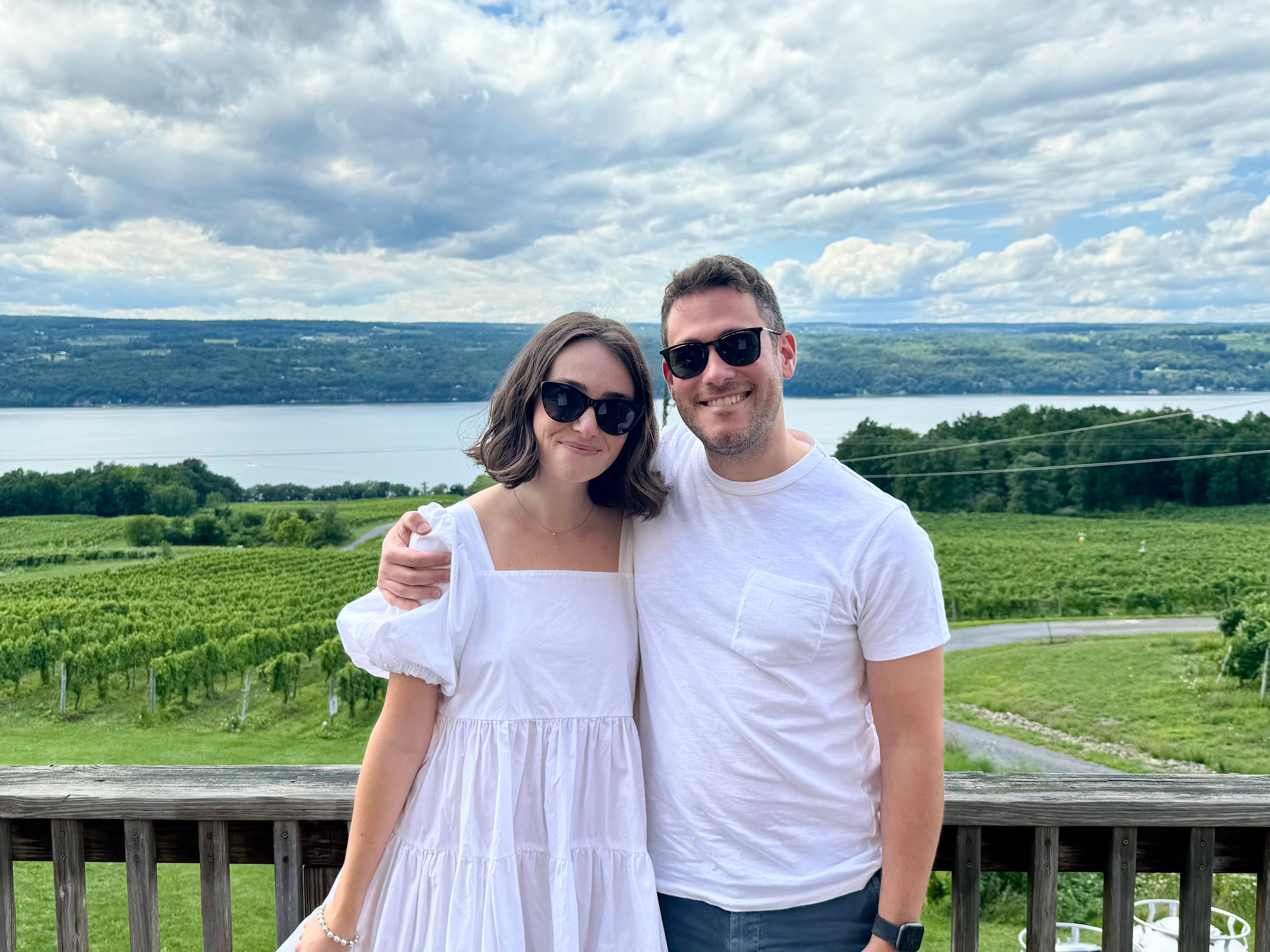 The author and her husband posing for a photo with Seneca Lake in the background. She's wearing a white puffed-sleeve dress and he's wearing a shite shirt, and they both have on sunglasses.
